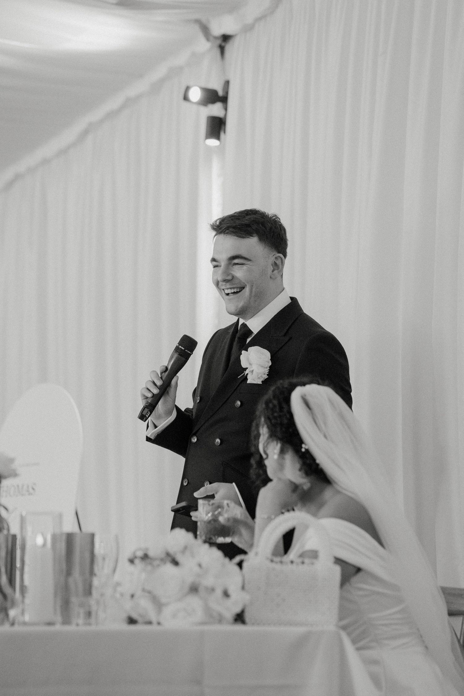 Groom laughs while giving wedding speech into microphone as bride sits beside him at reception table.