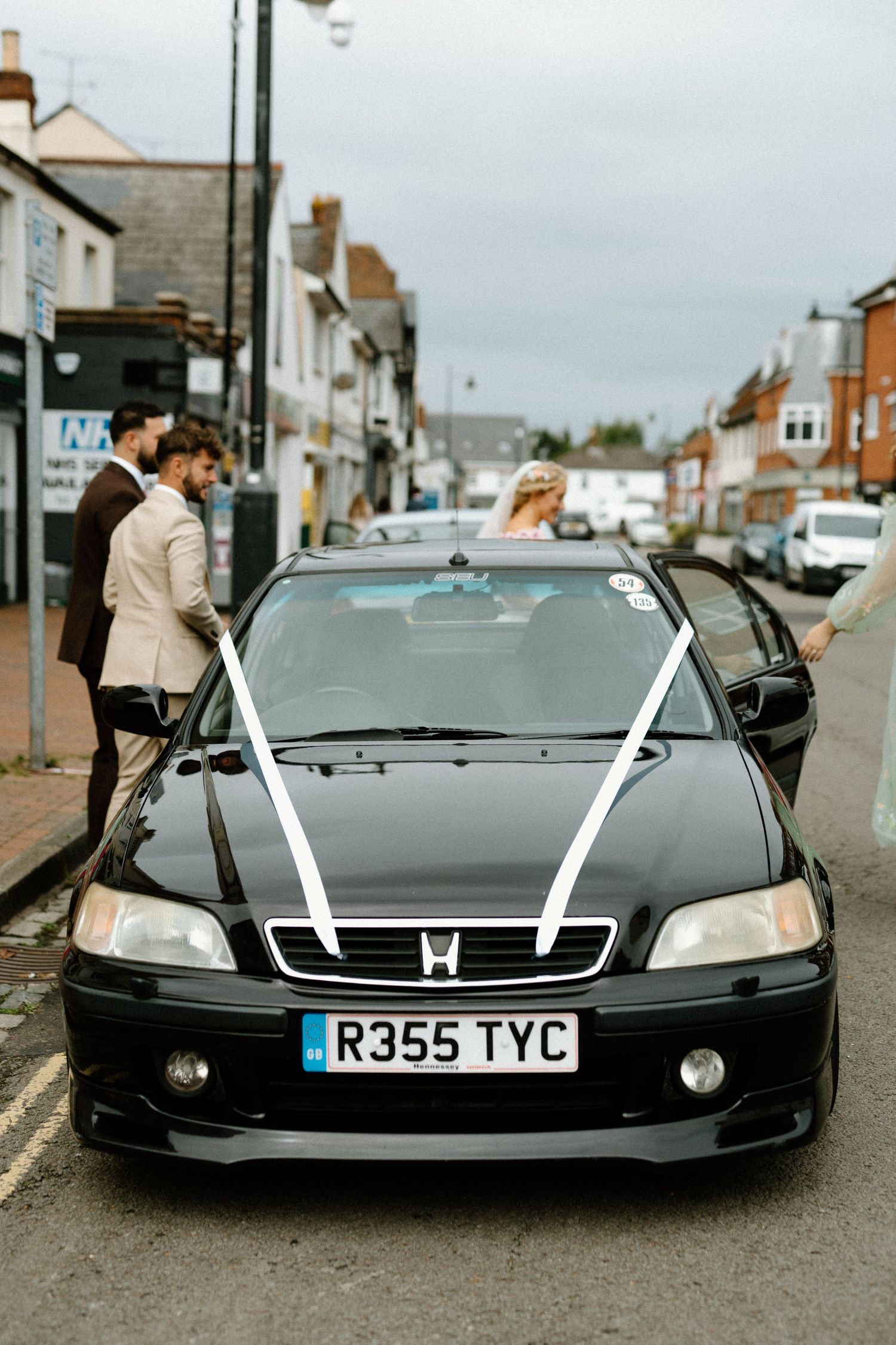 Bride and groom prepare to depart in a decorated black Honda wedding car on a town street.