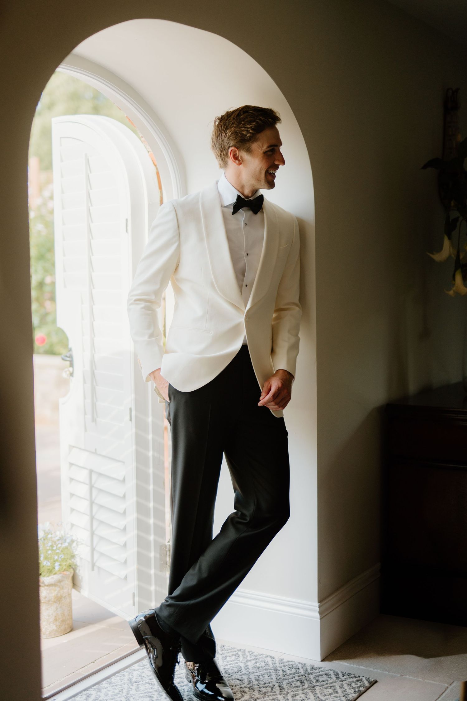 Stylish groom in ivory tuxedo and black bow tie leans against arched doorway smiling at wedding.