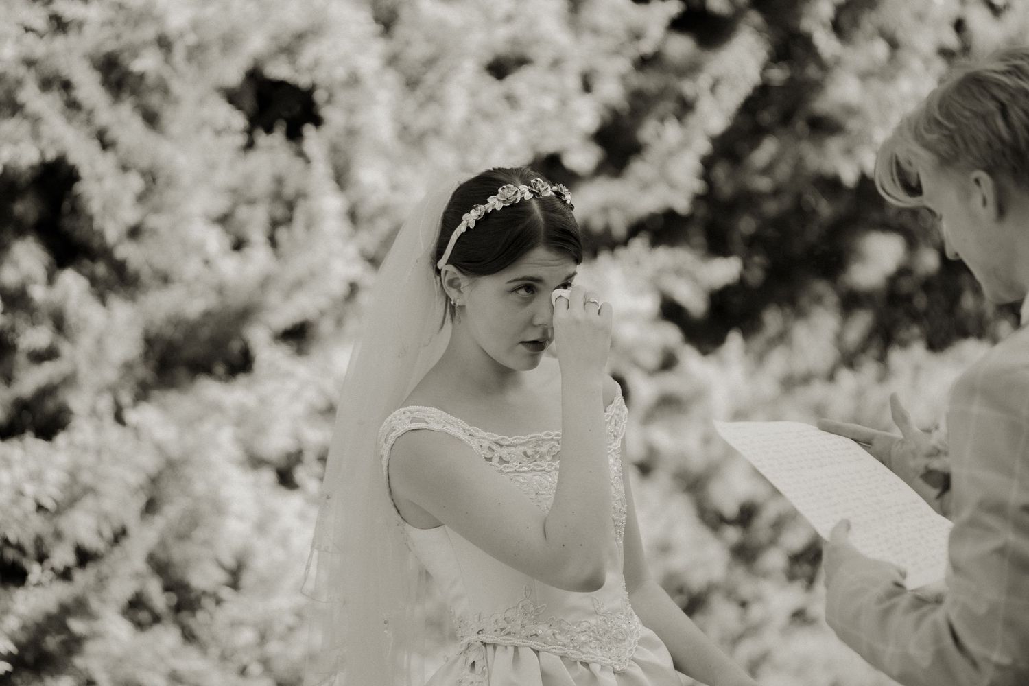 Black and white photo of tearful bride in veil wiping eyes during heartfelt wedding ceremony.