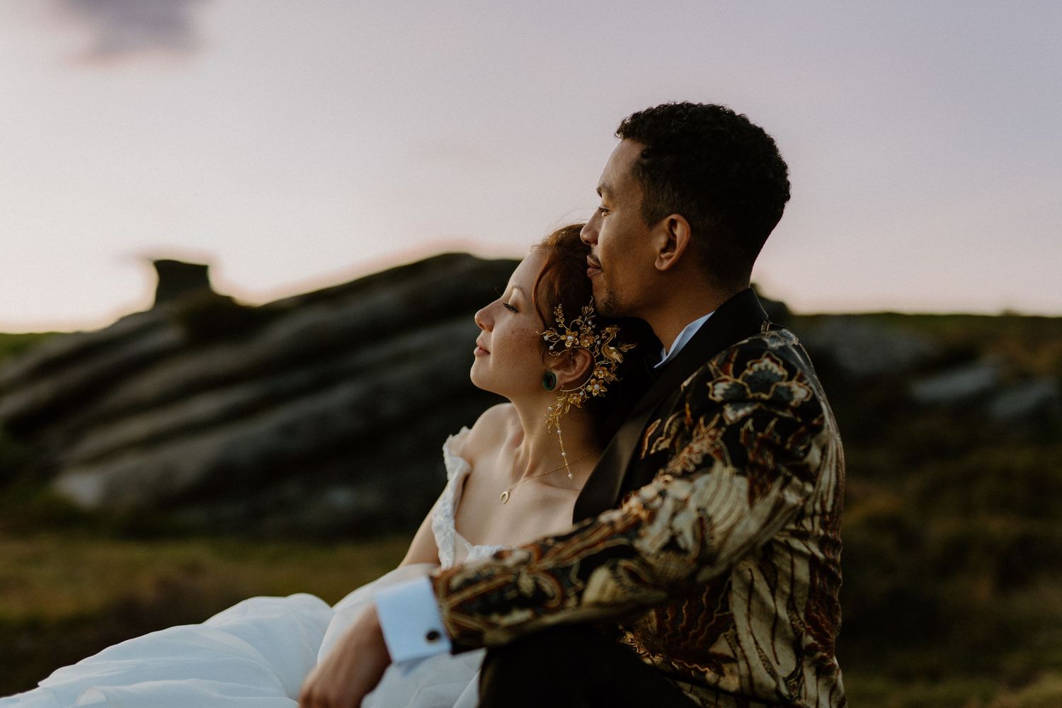 Couple sitting together at sunset on rocky moorland, groom kissing bride on the head.