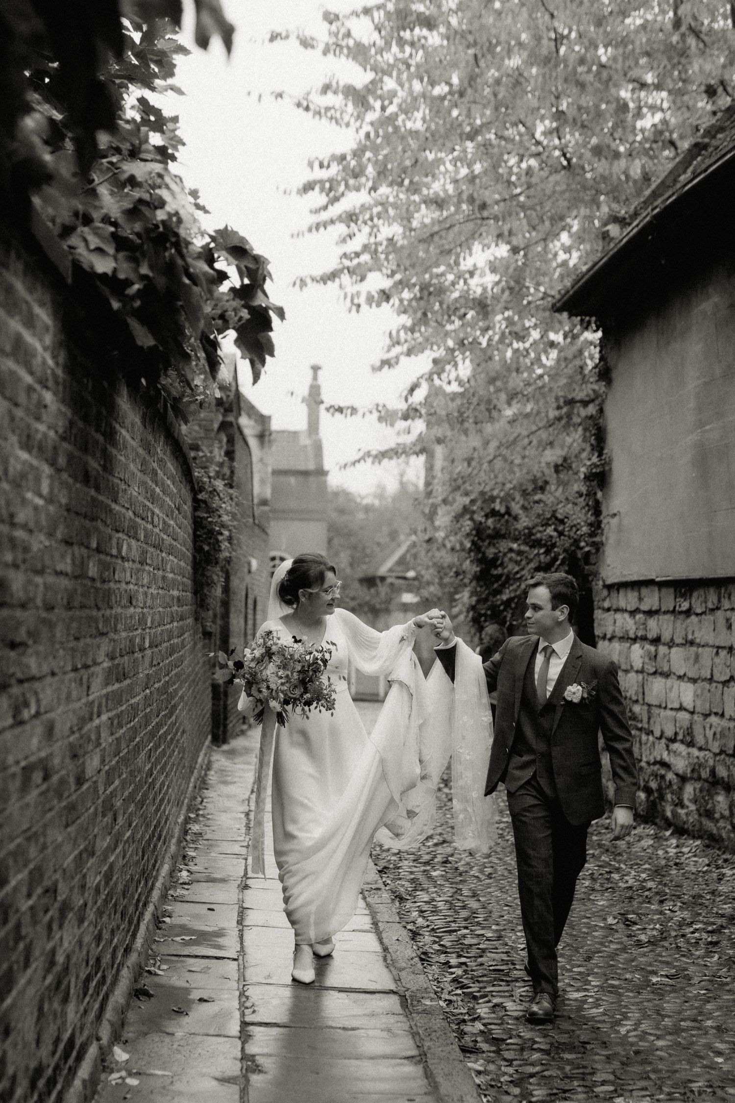 Bride and groom walk hand in hand down a charming cobblestone alley in black and white wedding photo.