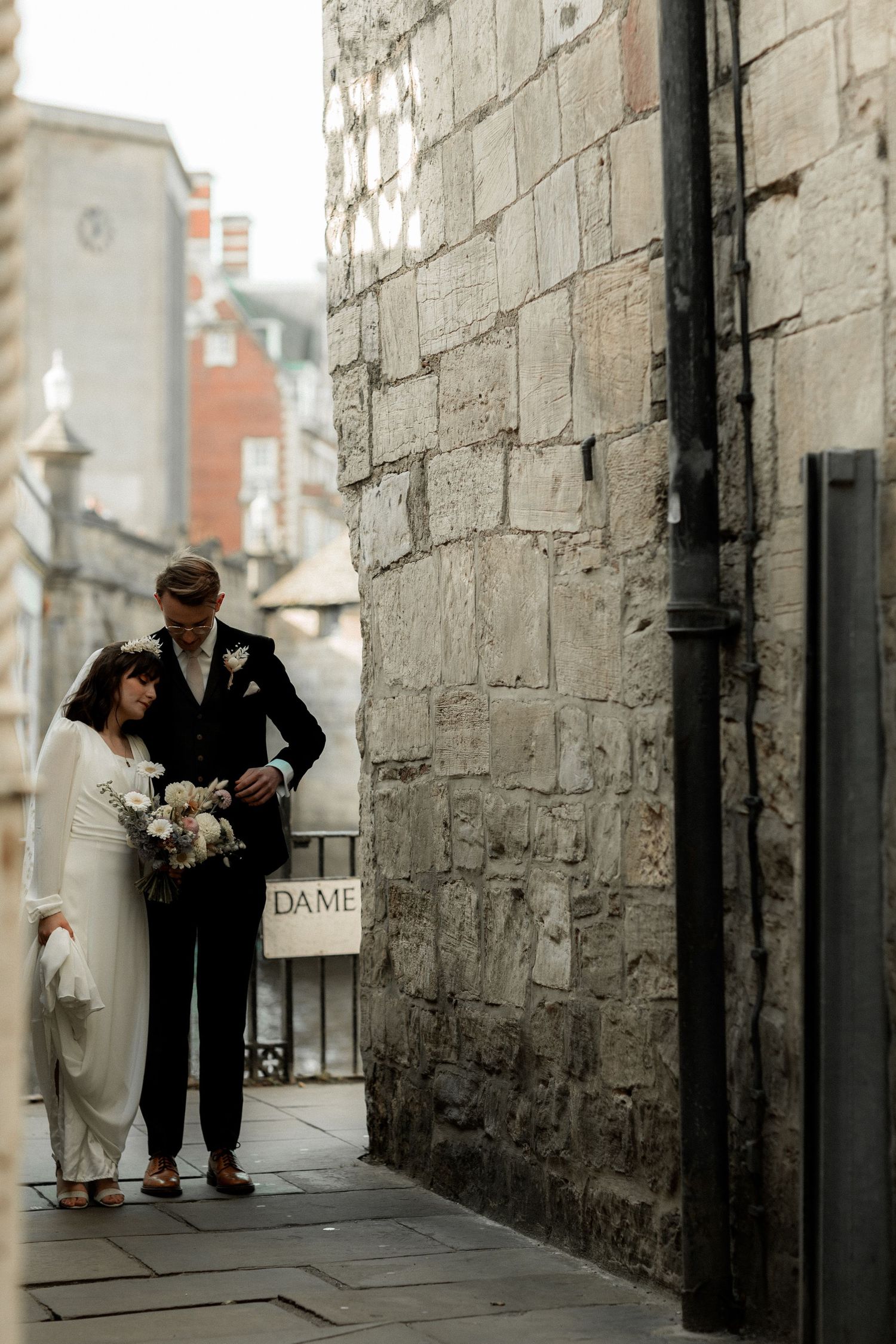 Wedding couple standing beside historic stone wall in urban setting.