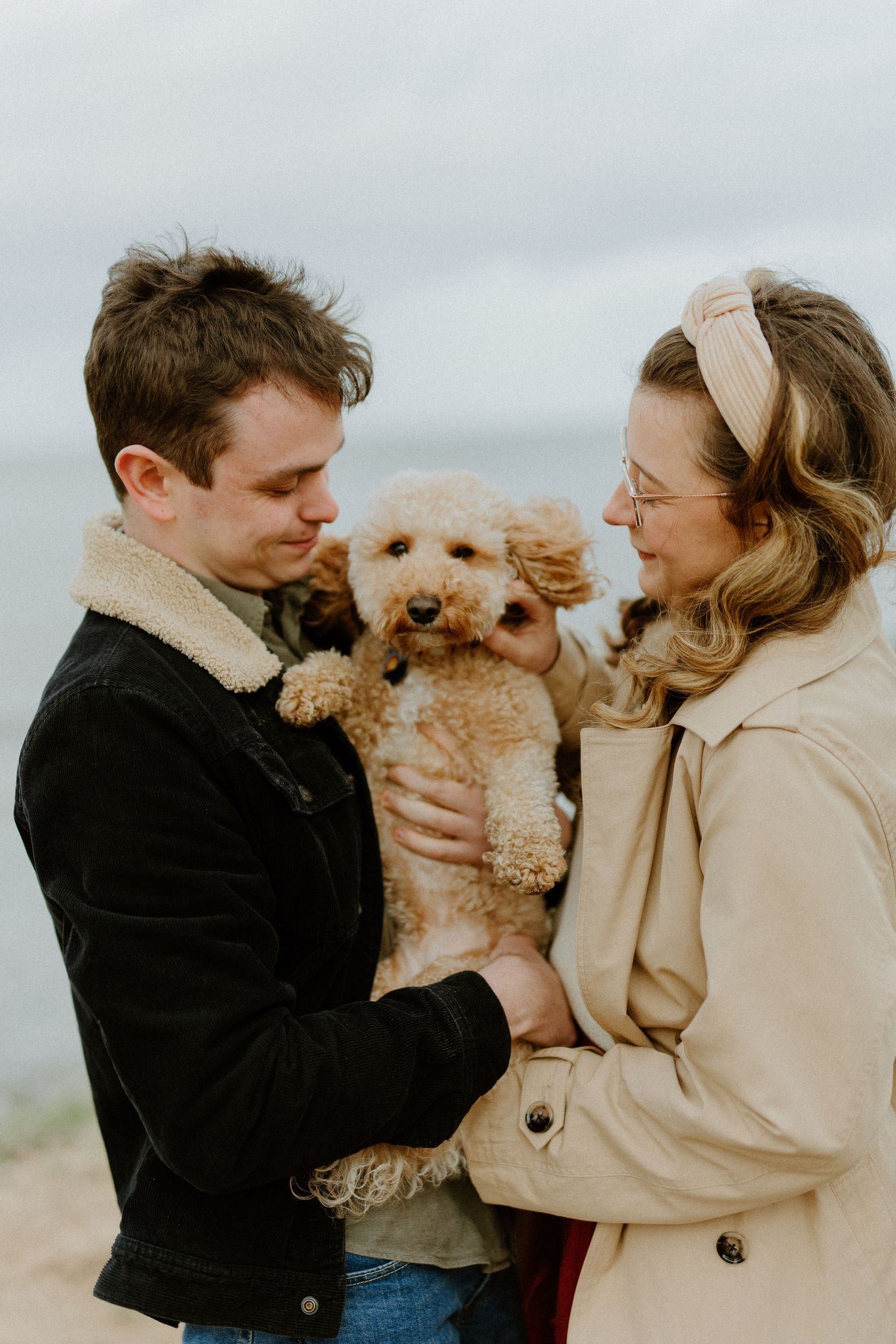A couple holds their adorable goldendoodle puppy outdoors on an overcast day, both wearing cozy winter coats.