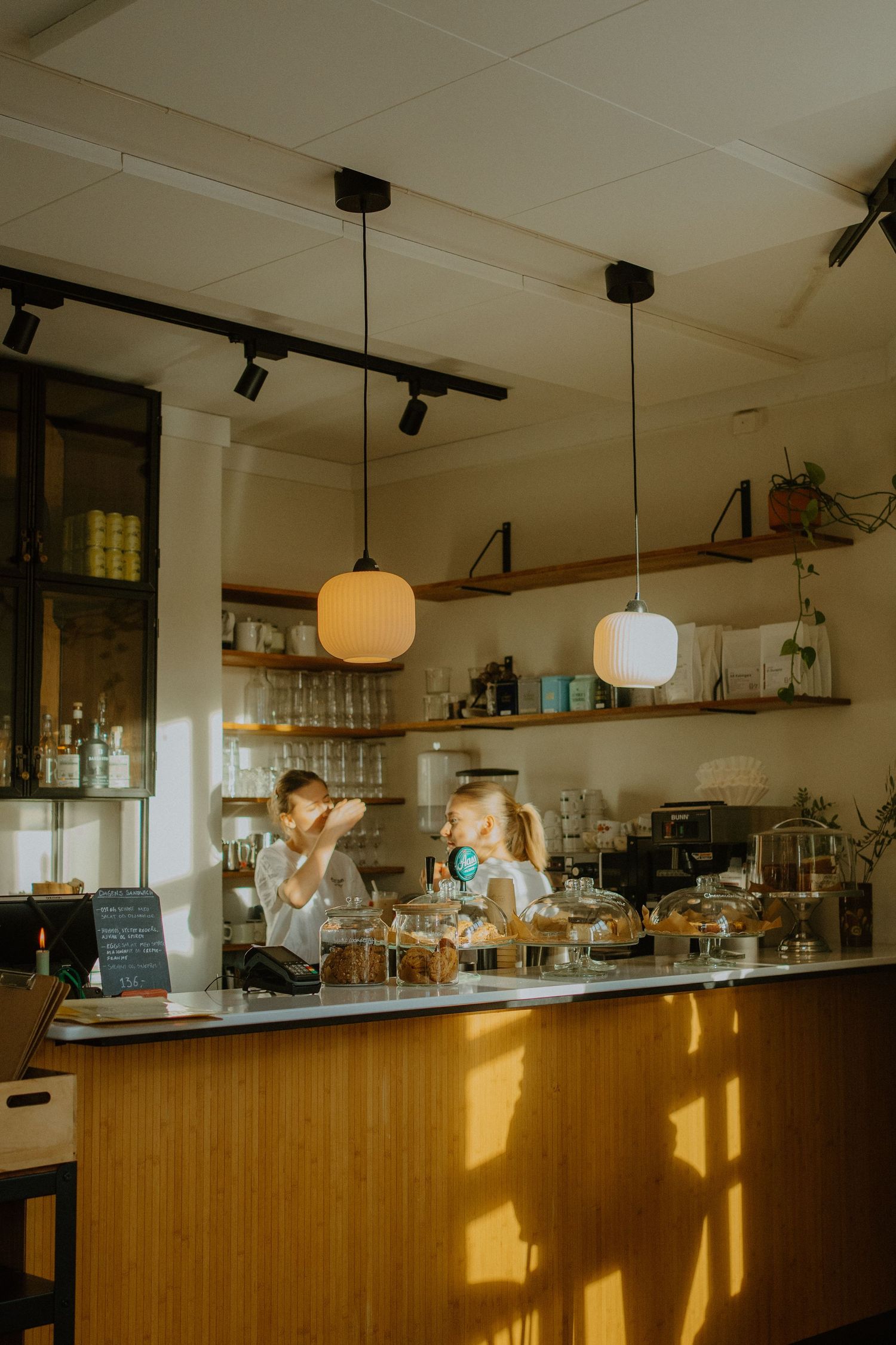 Modern cafe interior with wooden counter, pendant lights and rustic shelving displaying glassware and beverages.