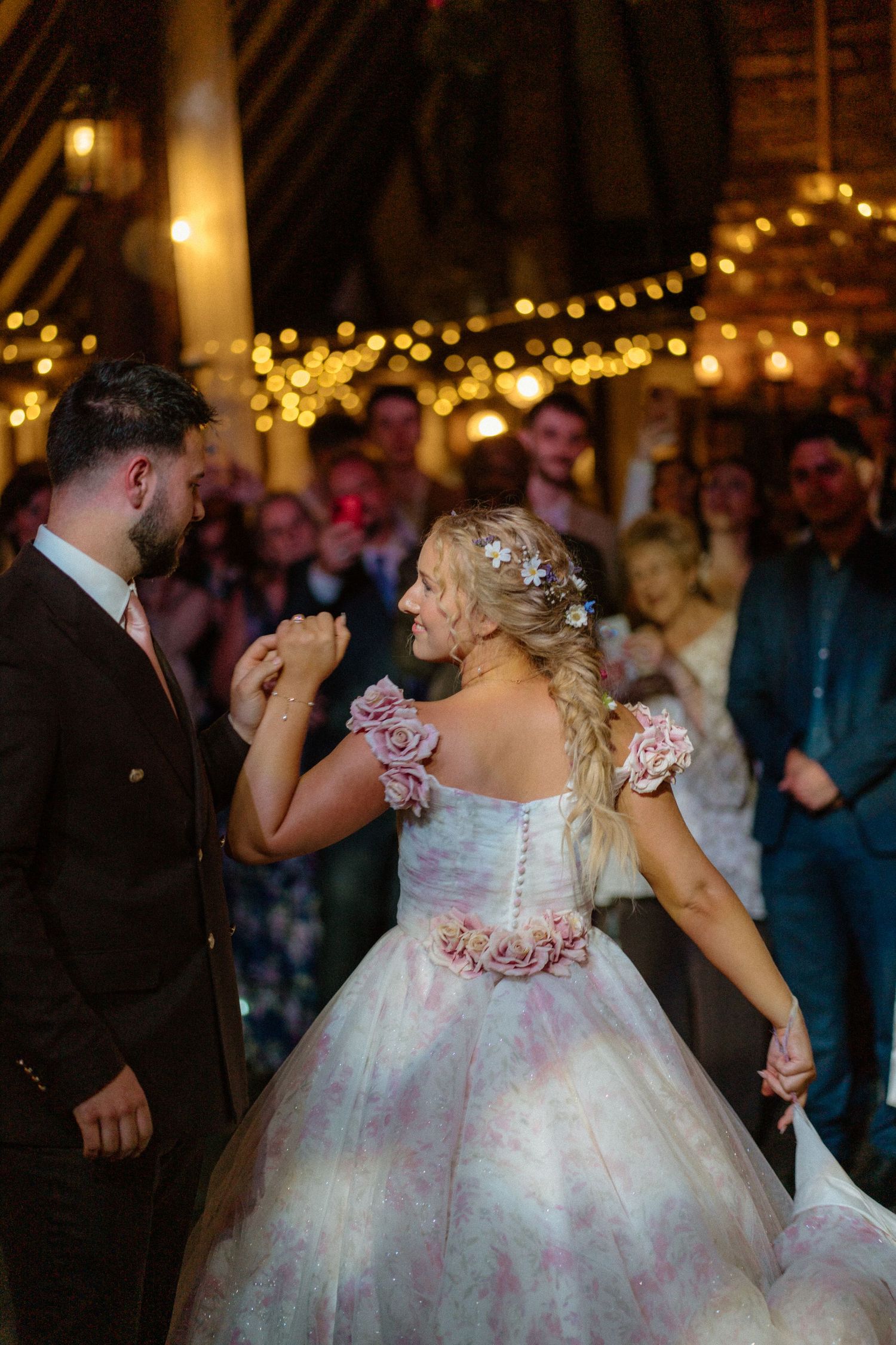 A couple dances at a wedding reception under twinkling string lights, with the bride wearing a floral pink and white gown.