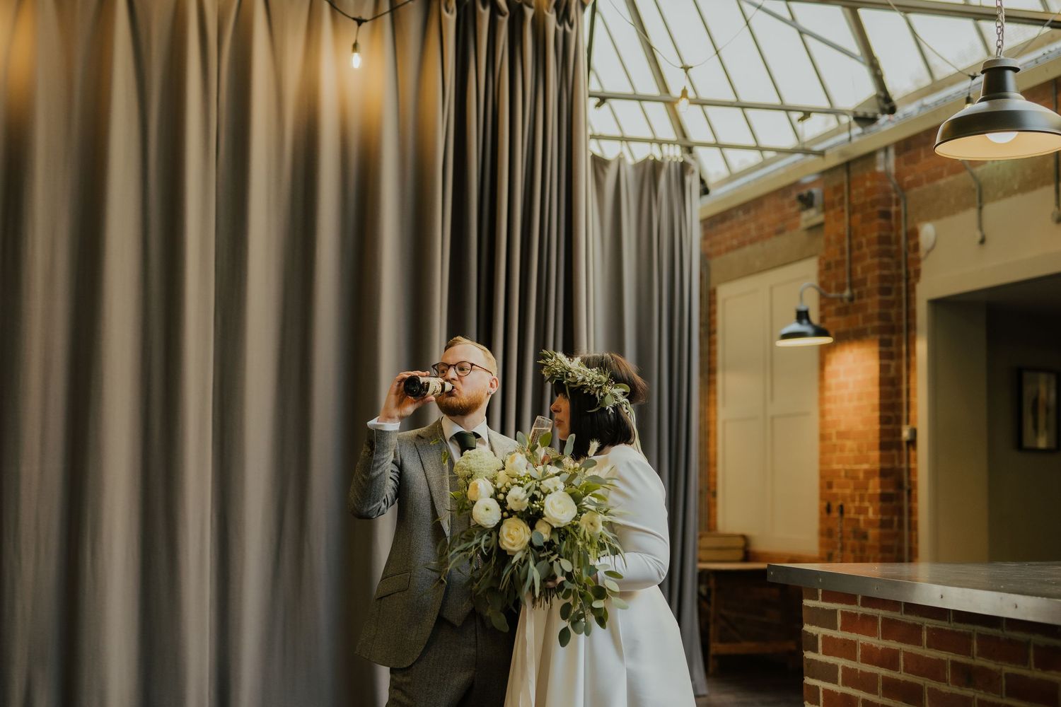 Bride with floral crown and groom in gray suit stand indoors near curtains in a rustic venue with exposed brick.