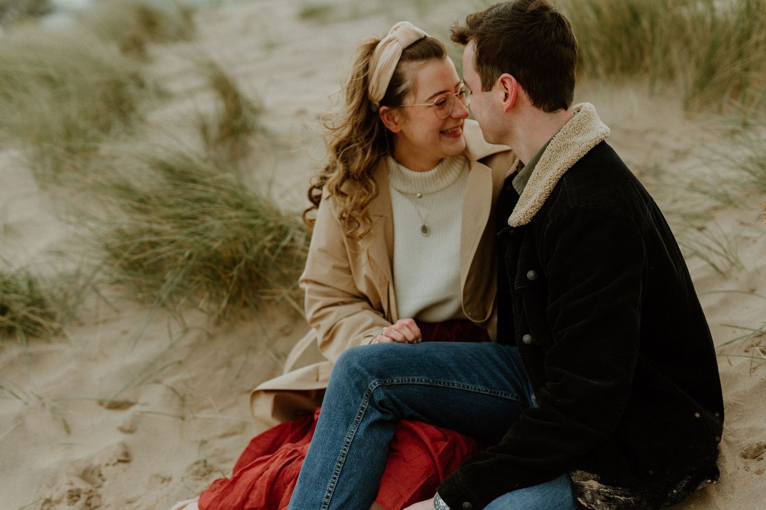 Couple sitting together on sandy dunes, sharing a romantic moment outdoors in casual autumn attire.