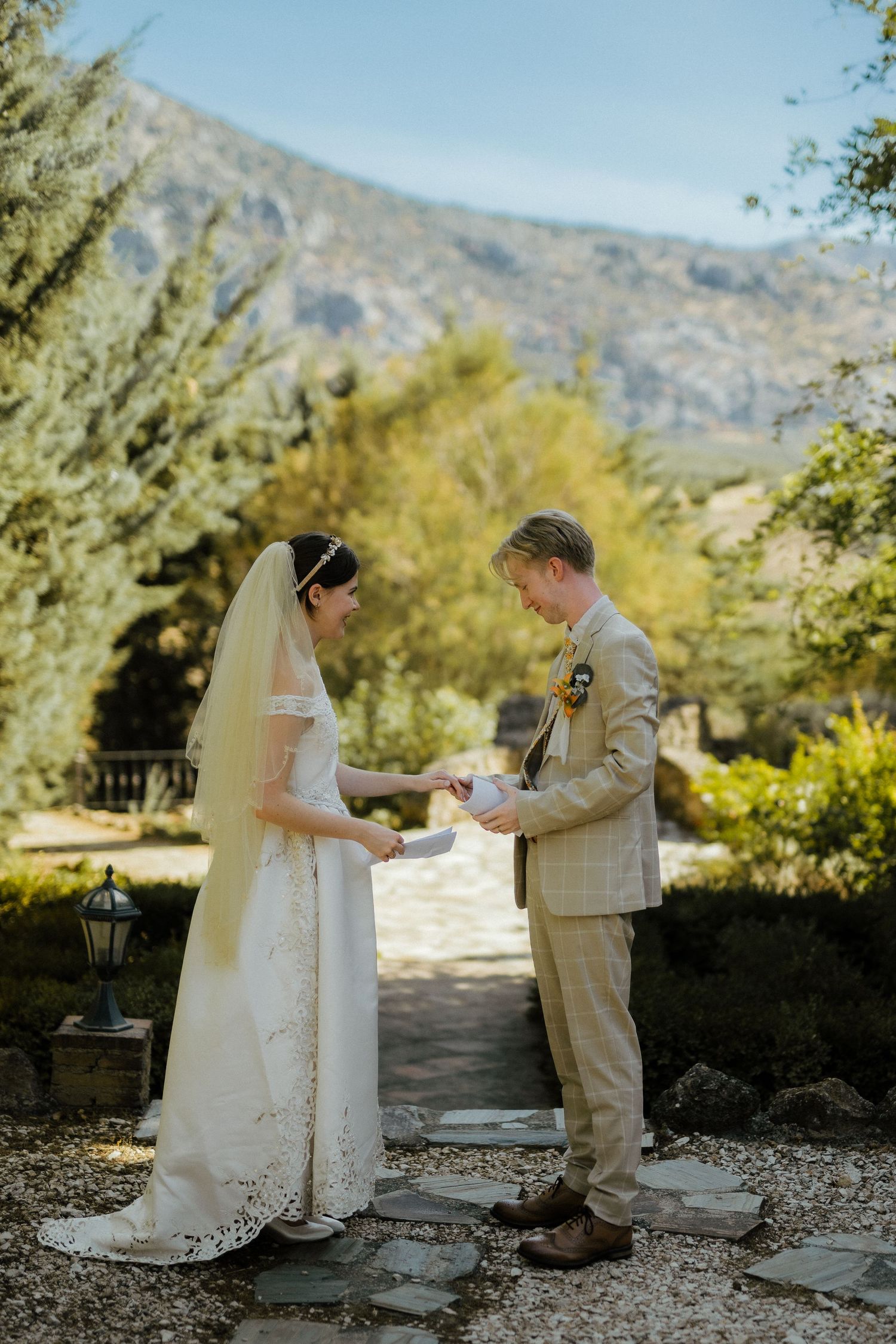 Bride and groom exchanging vows outdoors with mountain backdrop, bride in lace dress with veil.