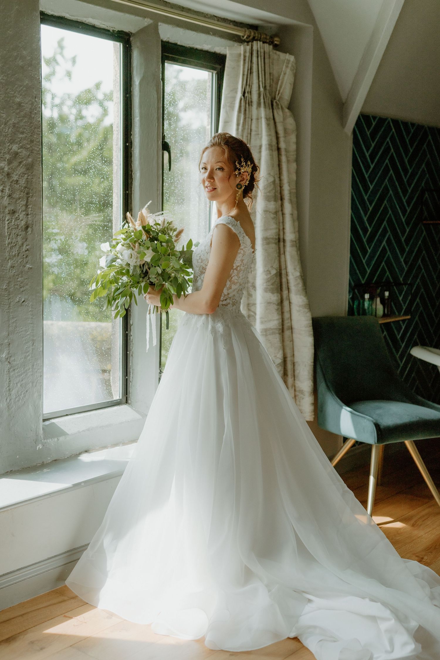 Elegant bride in white tulle gown holding bouquet while standing by a sunlit window.