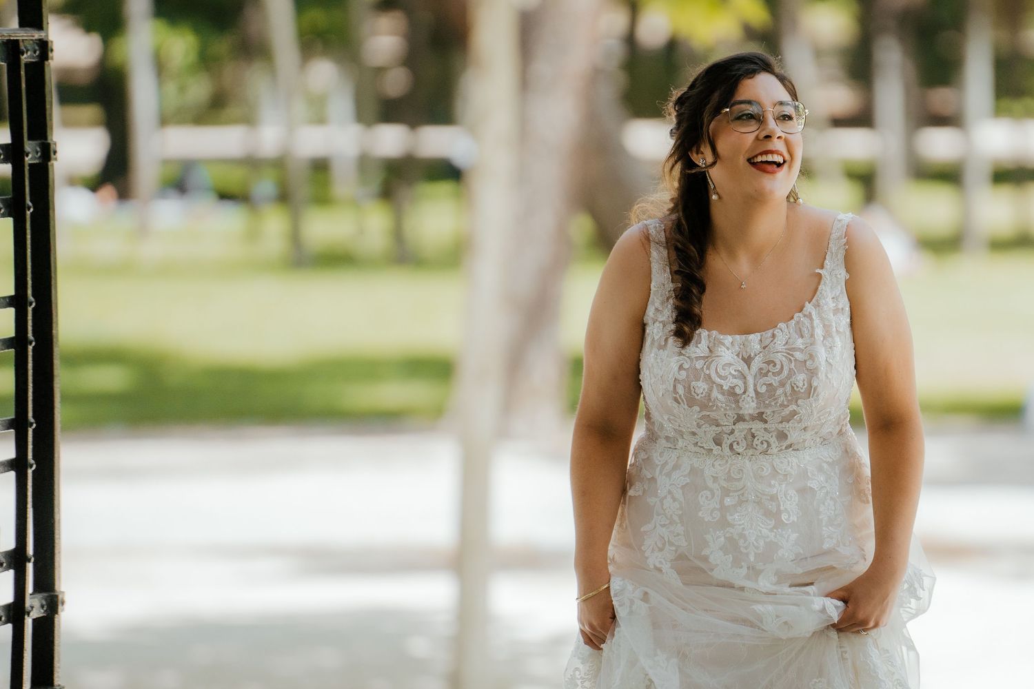 Joyful bride in lace wedding gown laughing outdoors in a sunlit garden before her ceremony.