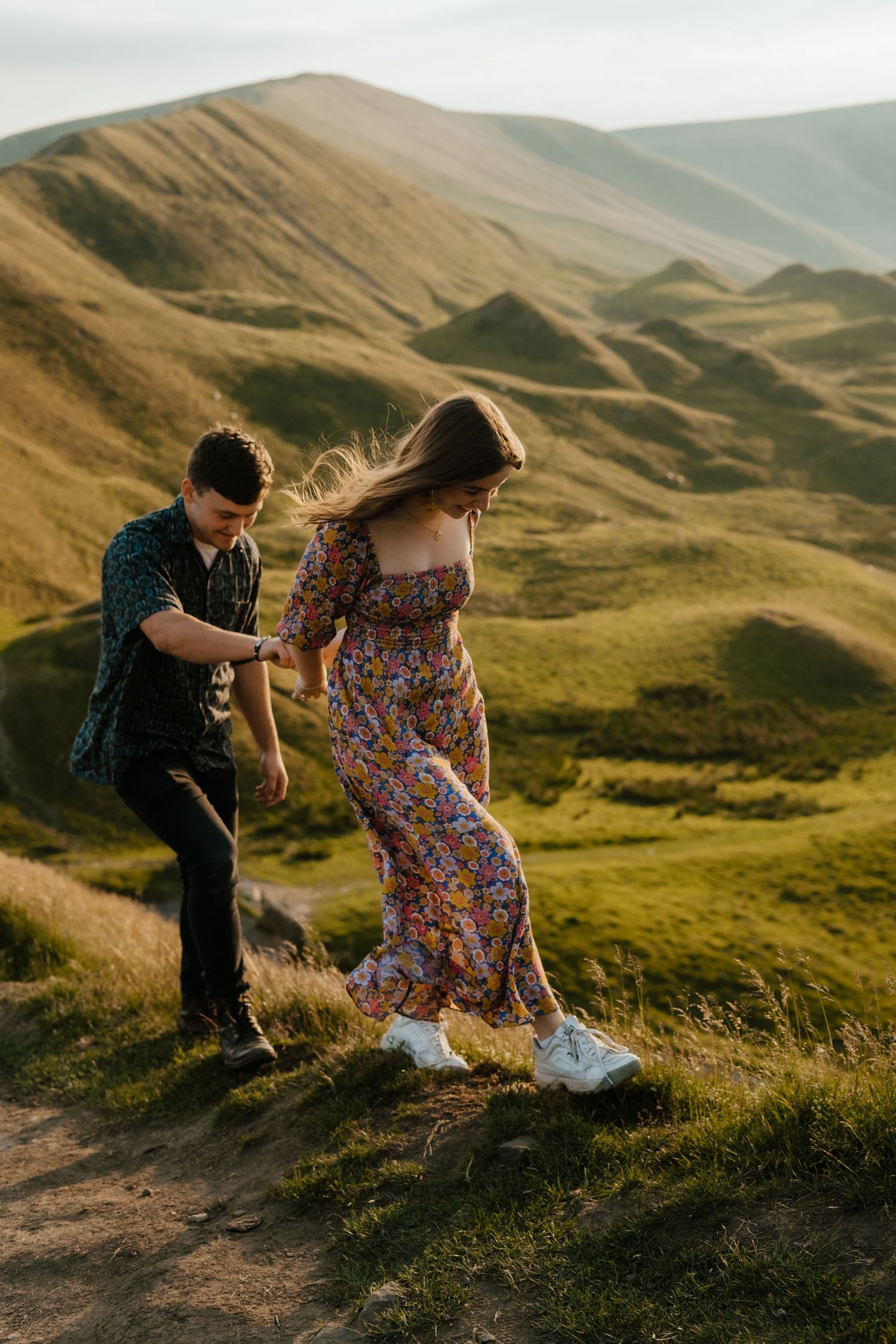Couple holding hands while walking up a scenic green hillside at golden hour.