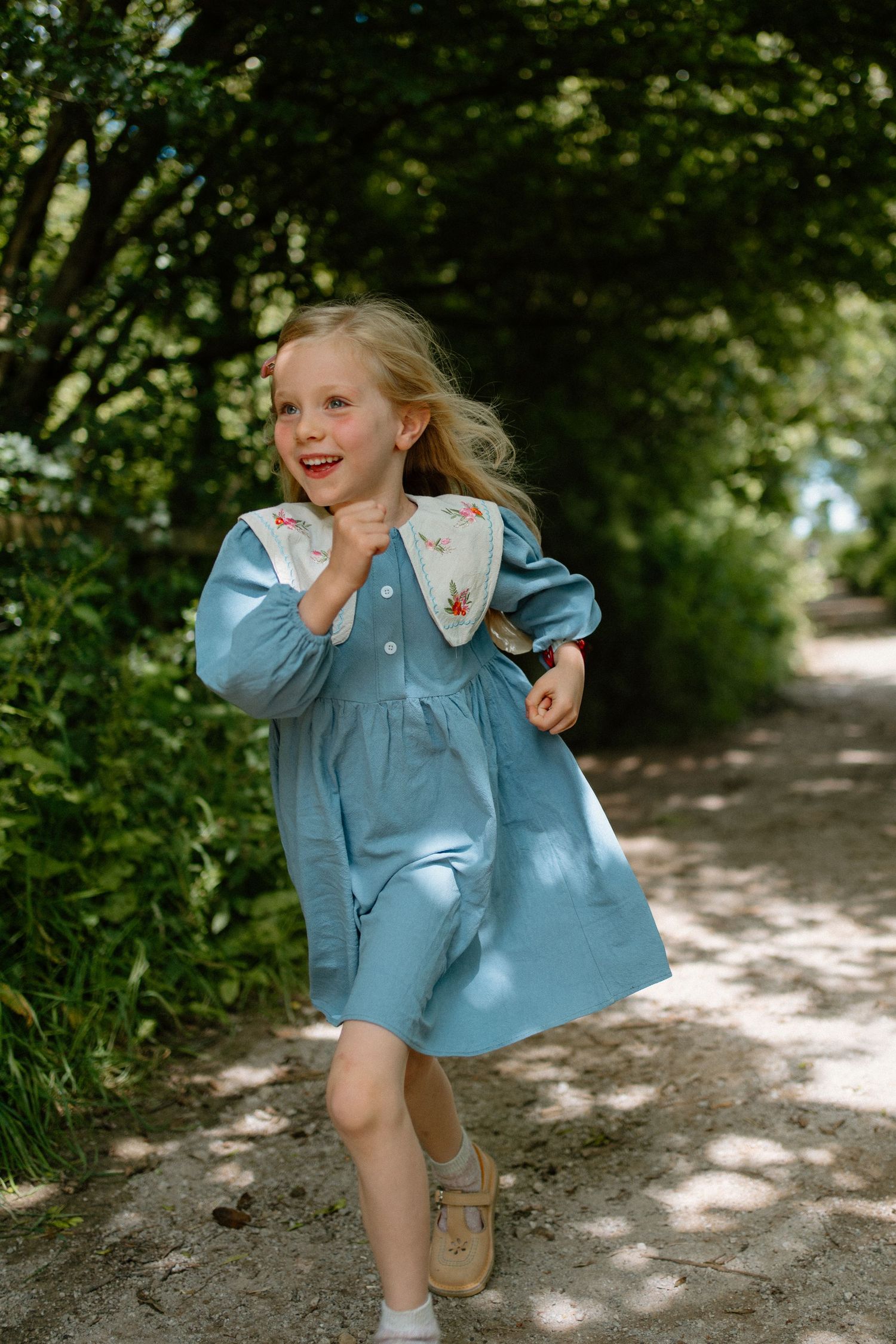 Joyful blonde girl in blue dress with embroidered collar running along a tree-lined garden path.