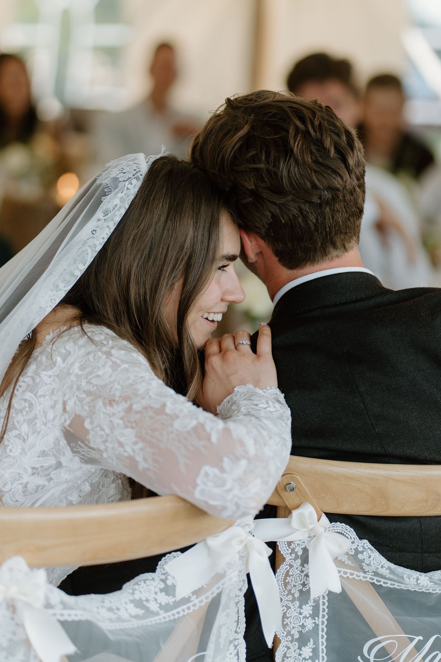 Bride in lace gown leaning close to groom during outdoor tented wedding reception.