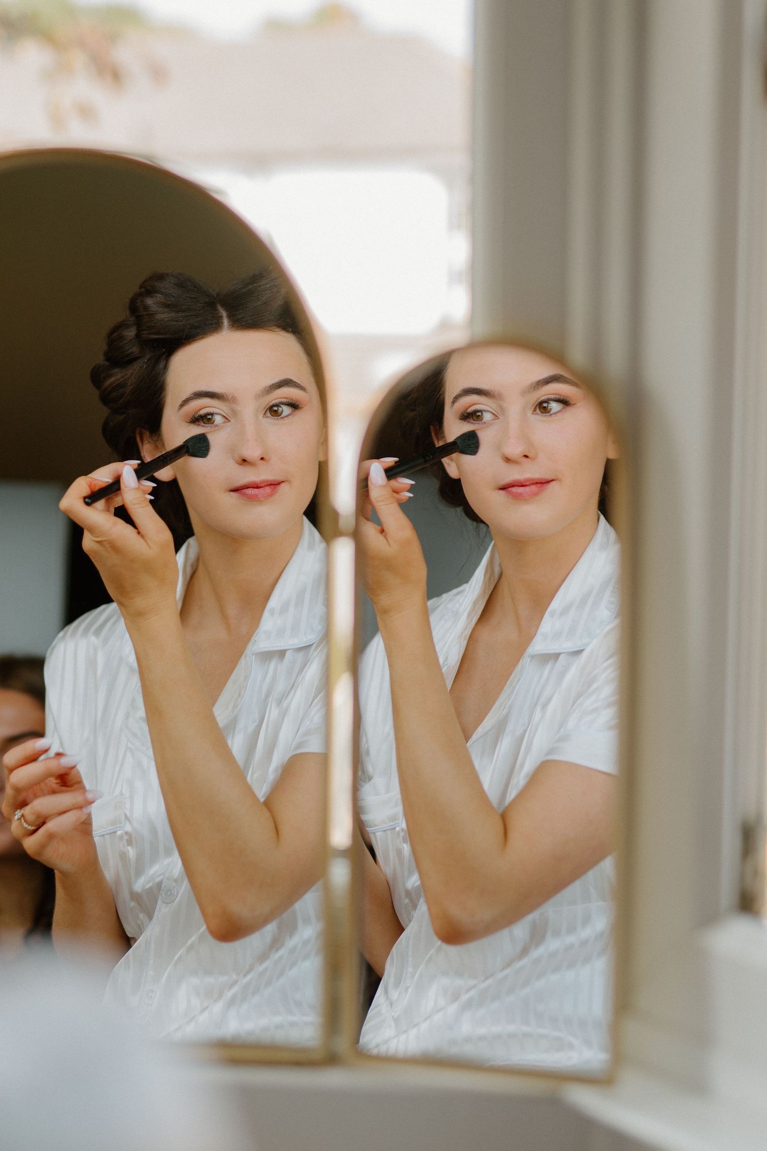 Bride applying makeup with brush while looking in mirror during wedding morning preparations.