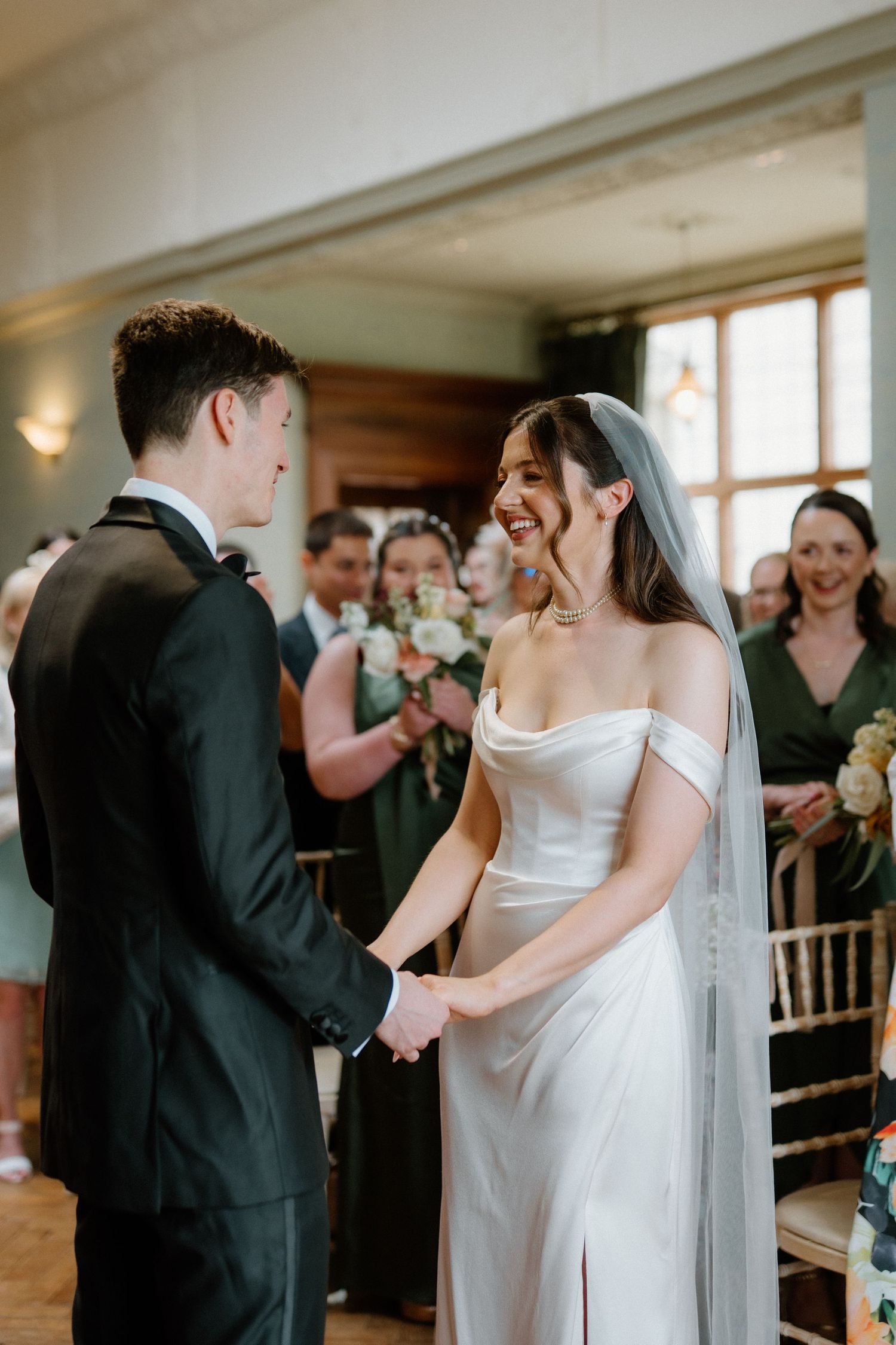 Bride and groom hold hands smiling during indoor wedding ceremony, bride wearing off-shoulder white gown and veil.