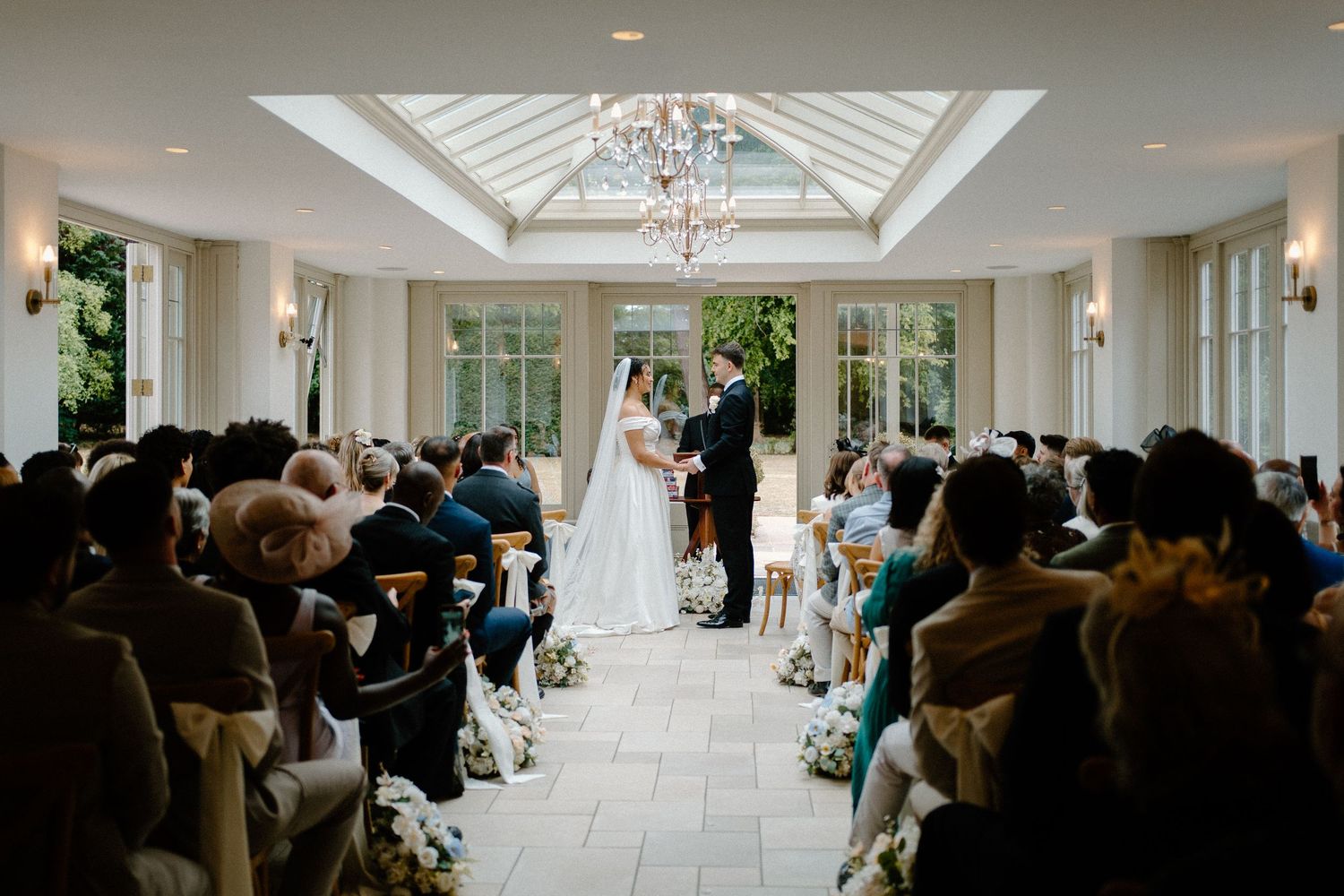 Elegant wedding ceremony in glass orangery with guests seated in rows.