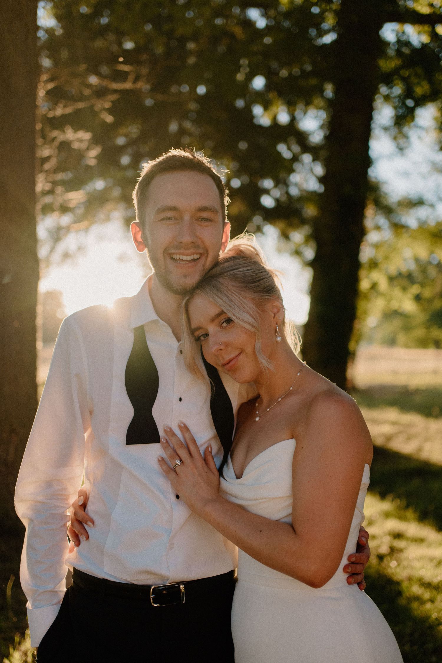 Smiling bride and groom embracing in warm golden sunset light among trees, bride showing off her wedding ring.
