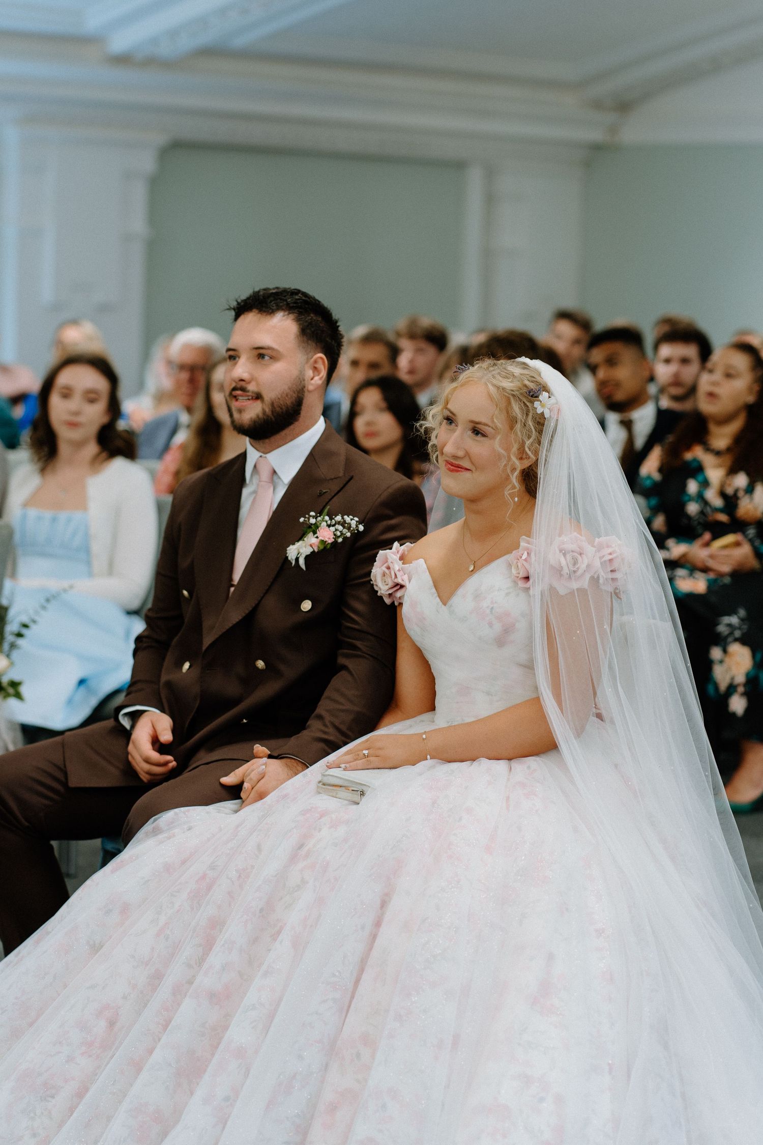 Bride and groom seated together during wedding ceremony, bride wearing pink floral ball gown with veil.