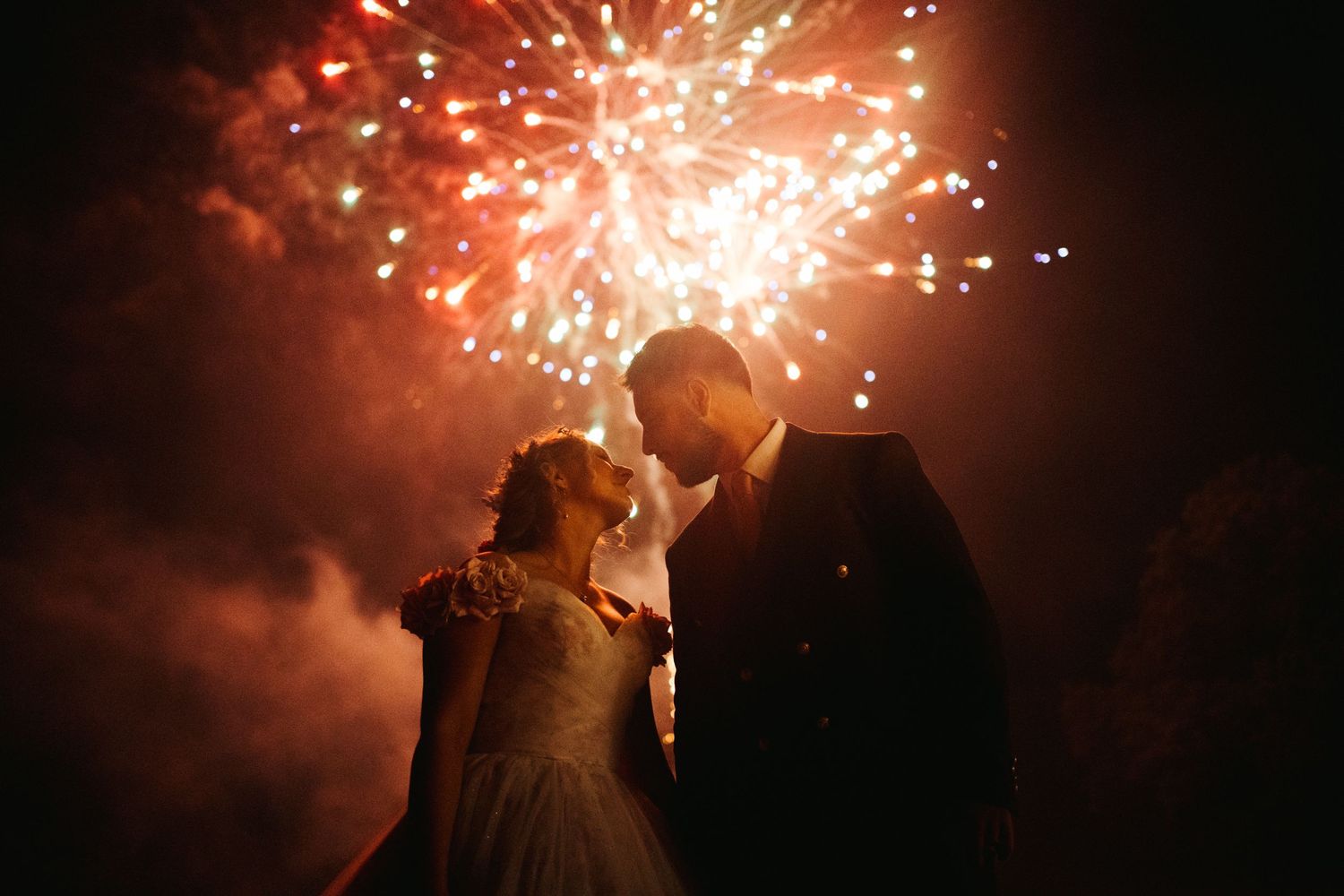 Bride and groom embracing under stunning red and gold fireworks display at their wedding celebration.