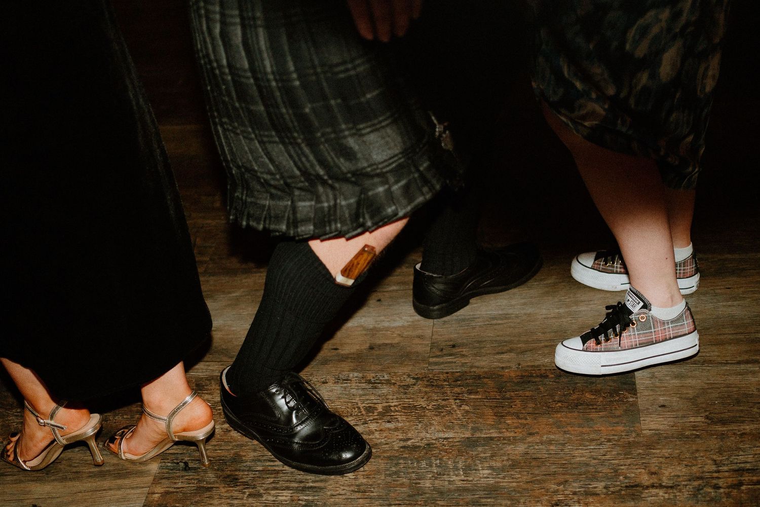 Close-up of wedding guests feet showing mix of dress shoes, Converse sneakers, and heels on wooden floor.