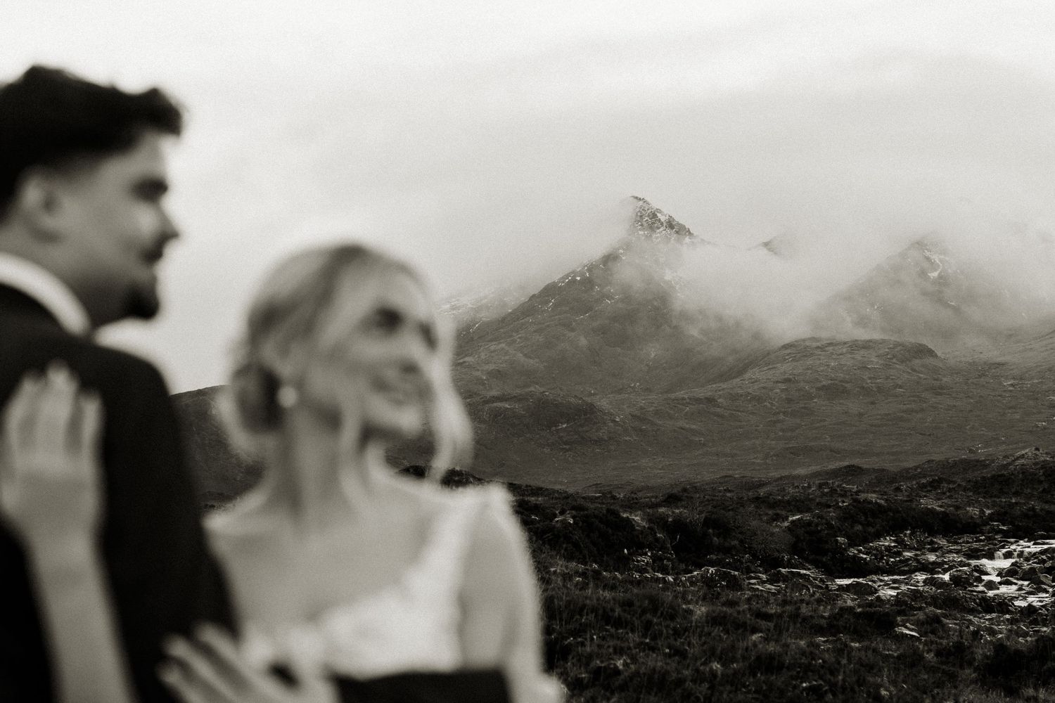 Black and white photo of bride and groom with misty mountain peaks in Scottish highland setting.
