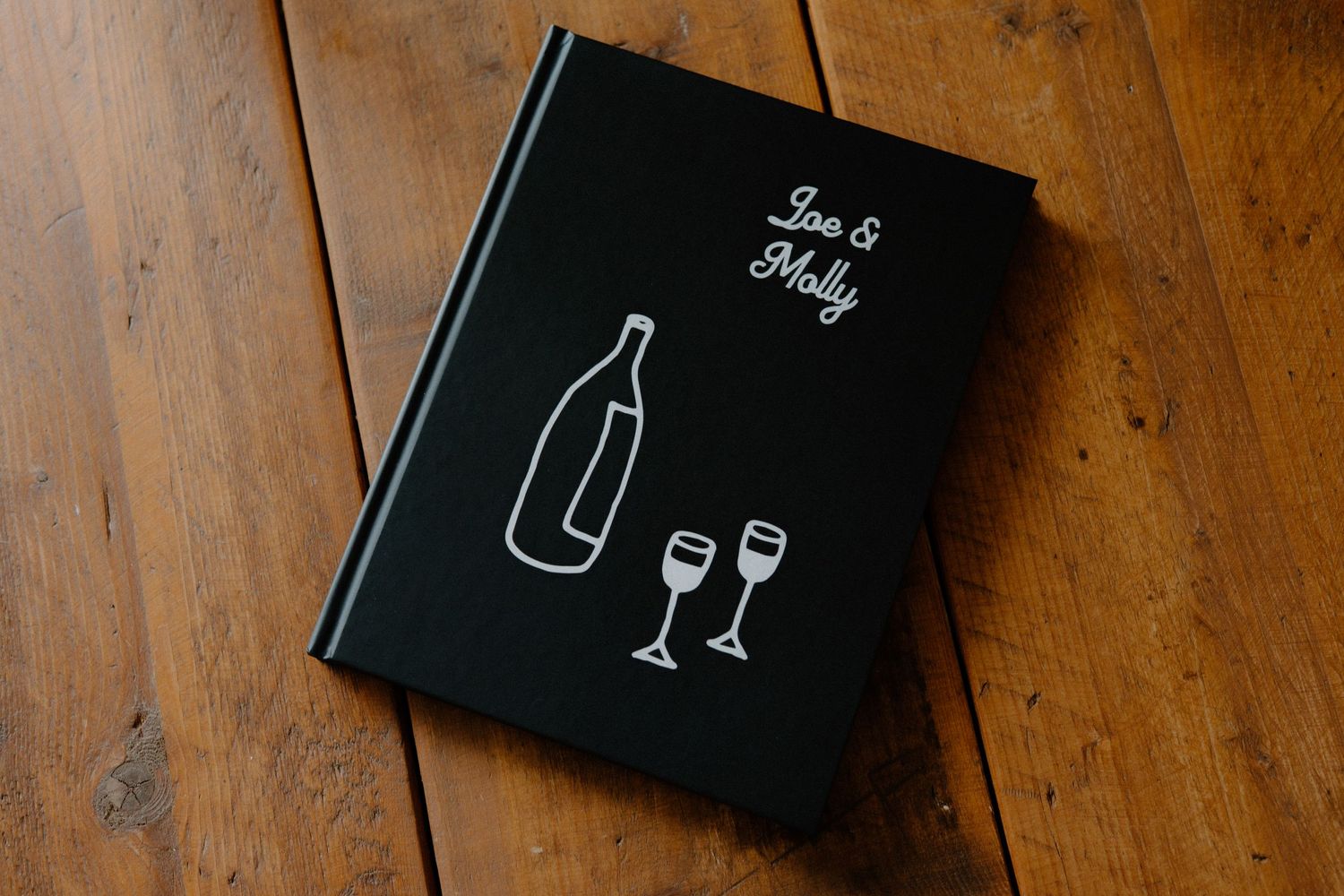 Black hardcover book with minimalist wine bottle and glasses illustration on wooden surface.