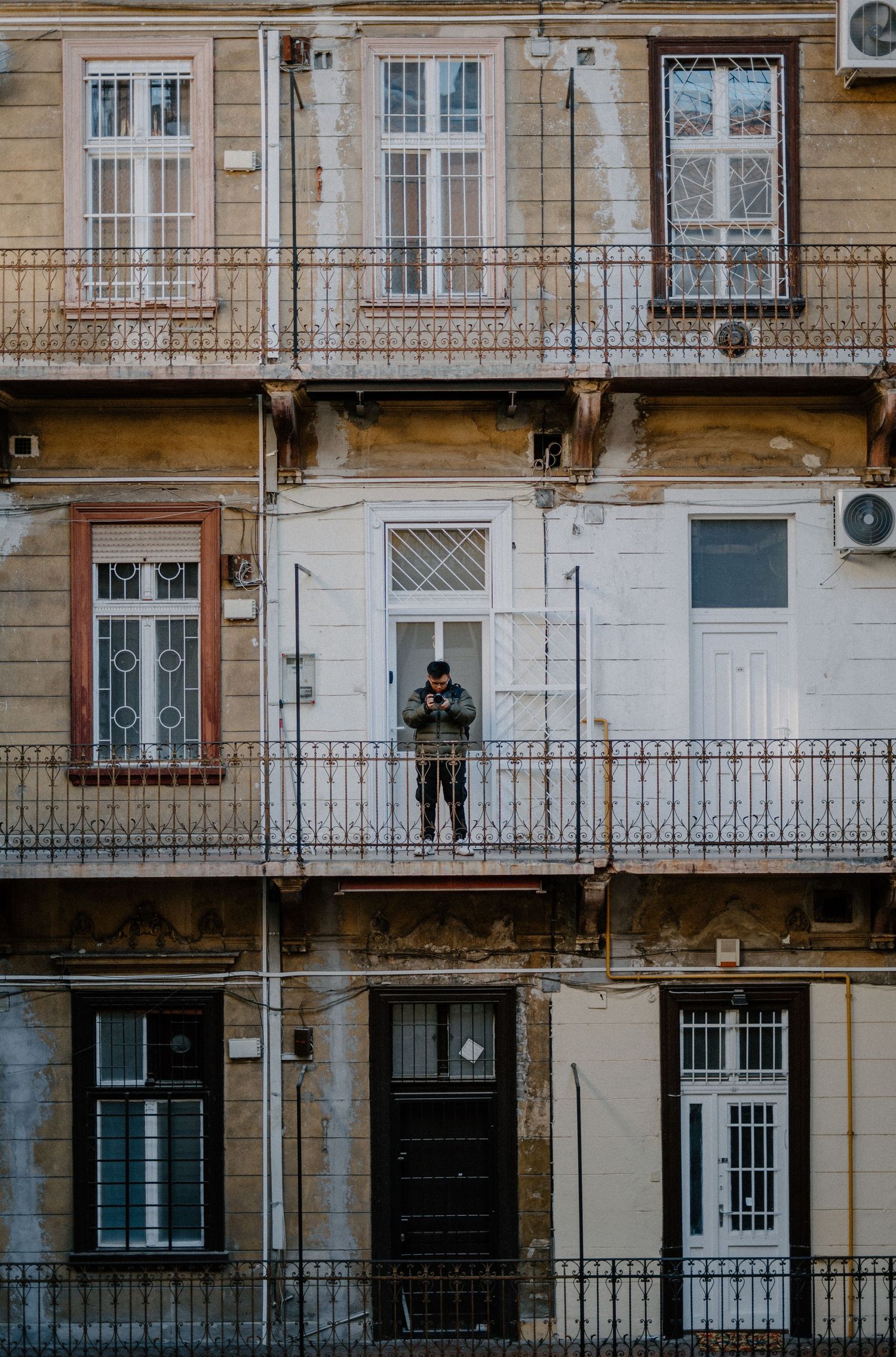 An old European apartment building facade with iron balconies and windows, showing a person standing on a middle floor balcony.