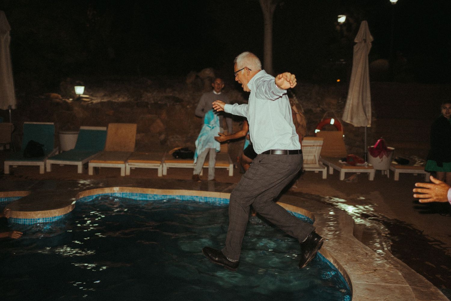 An older gentleman in formal attire jumping into an outdoor swimming pool at a wedding reception.