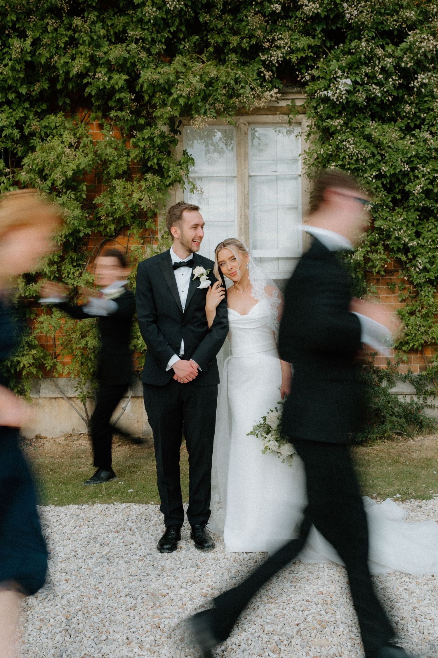 Bride and groom standing still as wedding guests blur past them in motion before an ivy-covered brick wall.