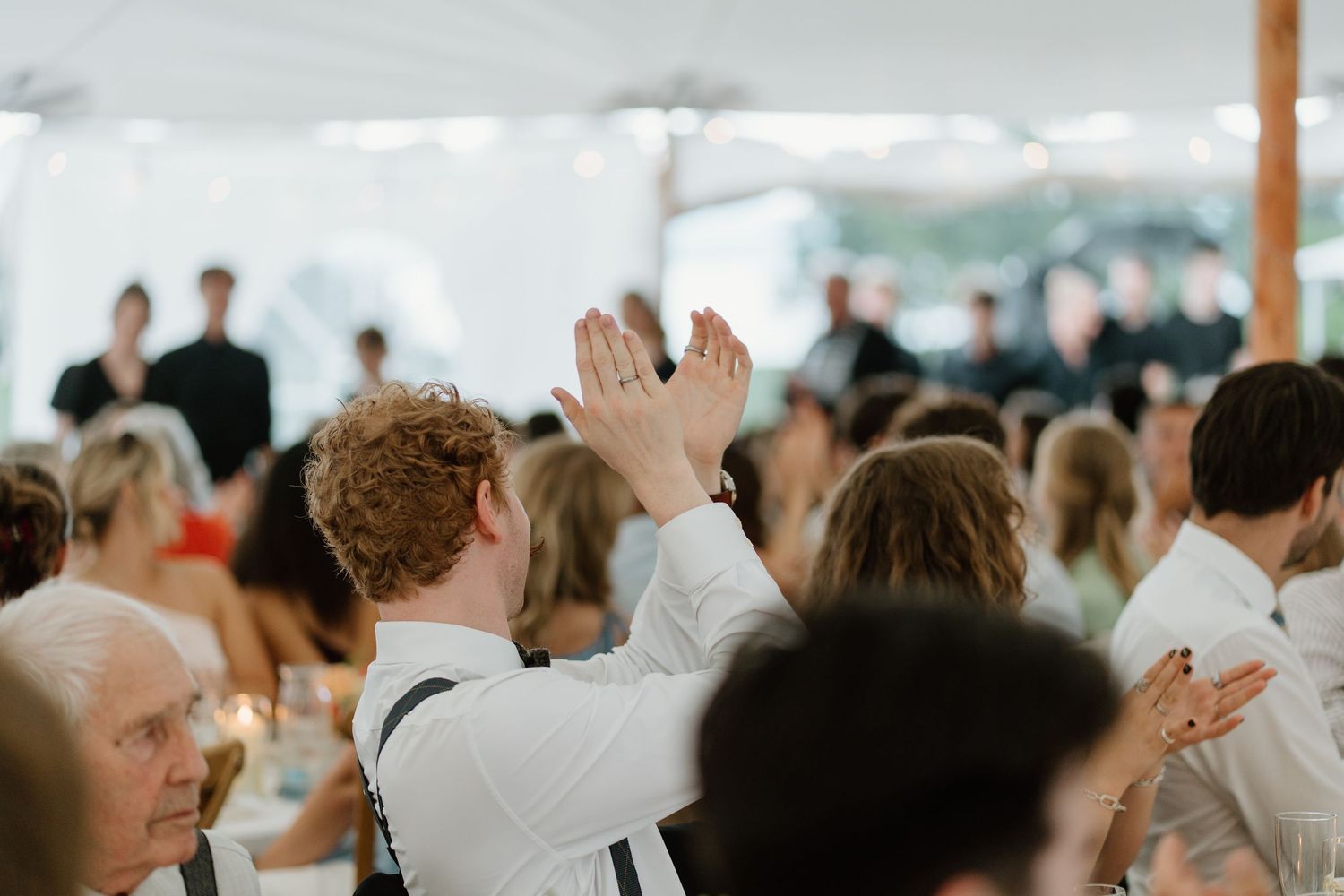 Wedding guests clap and celebrate during lively reception under a white tent with string lights.