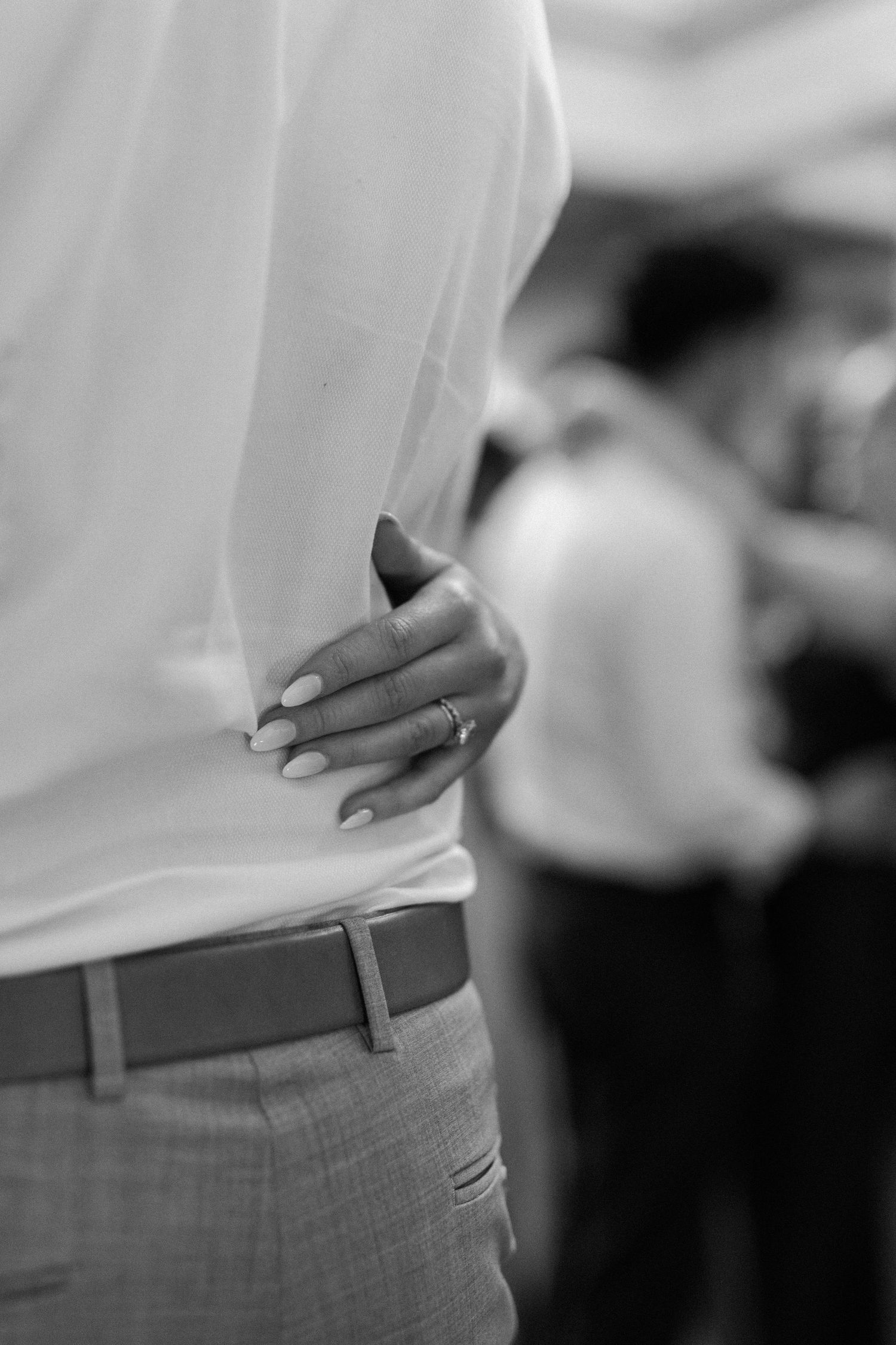 Black and white close-up of a bride's hand with ring resting on groom's back at wedding.