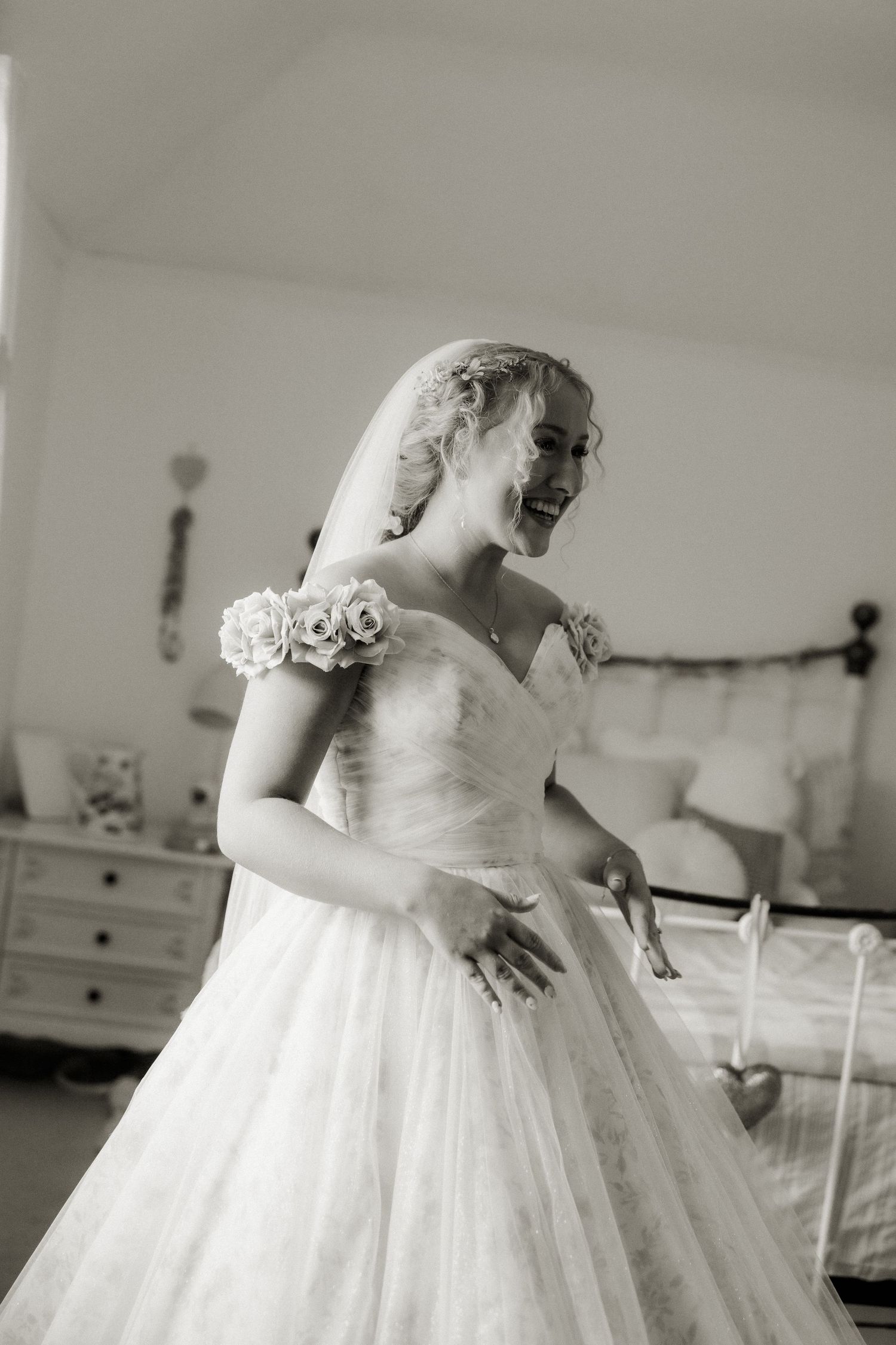 Black and white photo of smiling bride in rose-adorned ball gown twirling in bedroom before wedding.