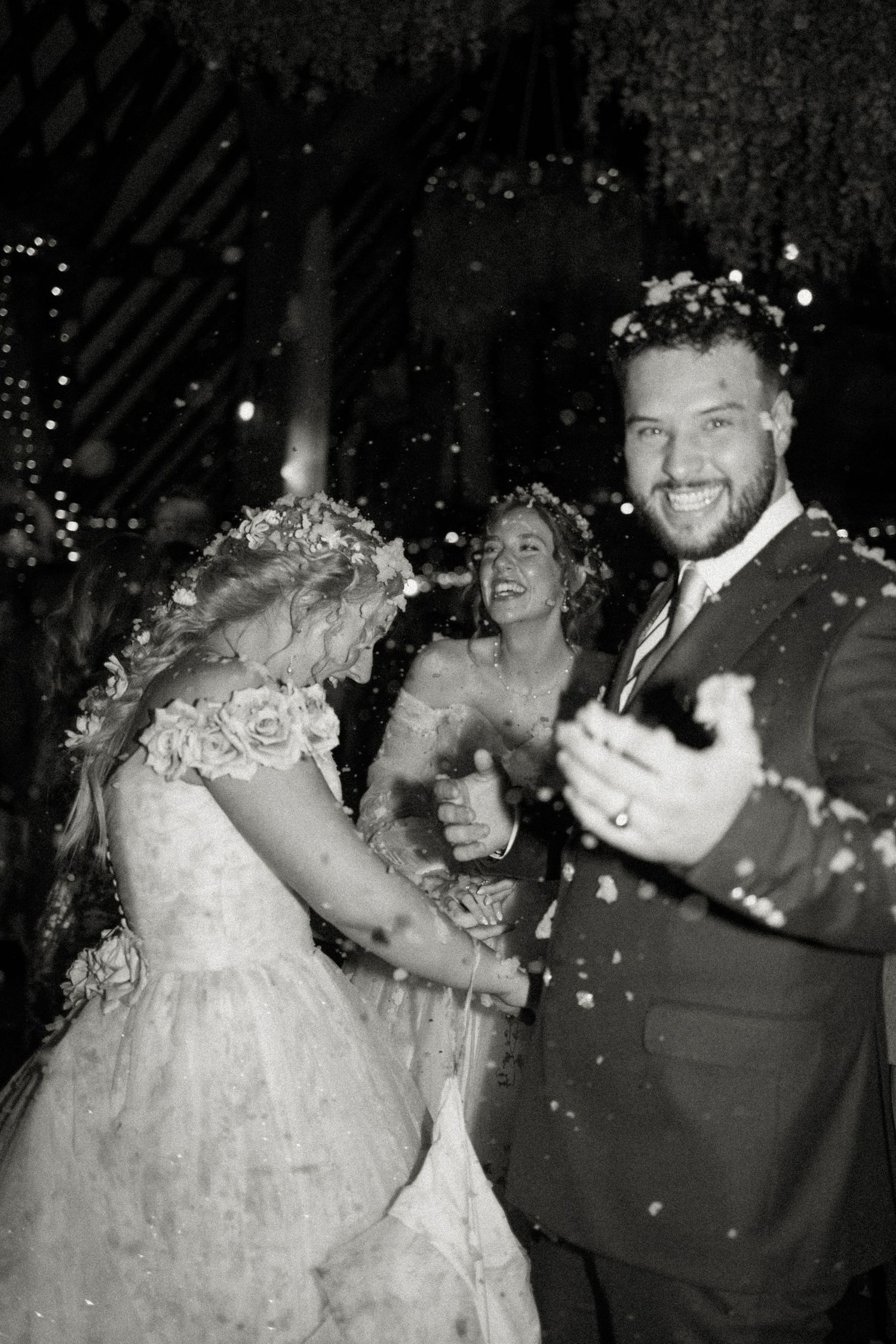 Black and white photo of smiling bride and groom dancing in confetti shower with guests celebrating.