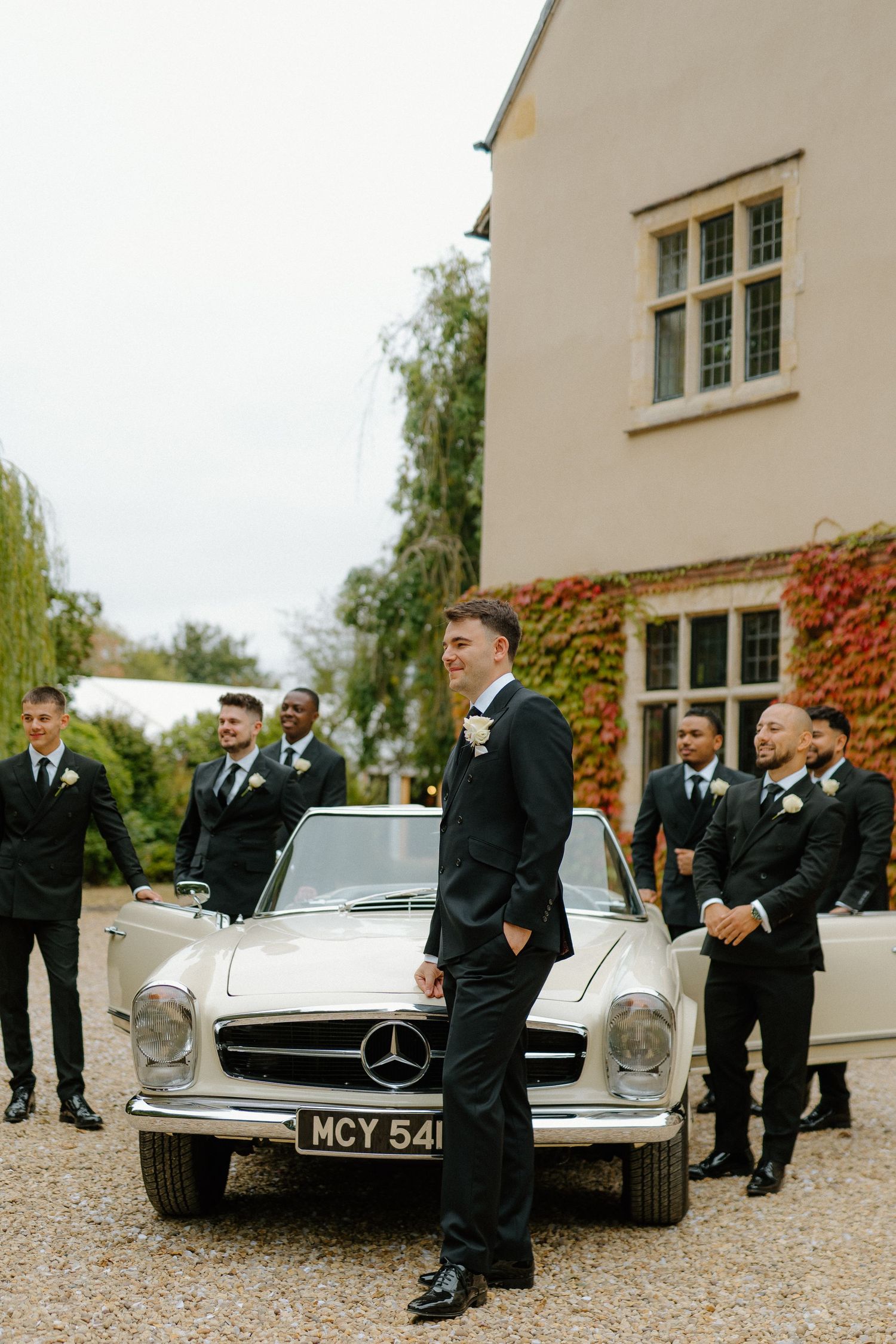 Groom and groomsmen in black suits posing with vintage Mercedes wedding car.