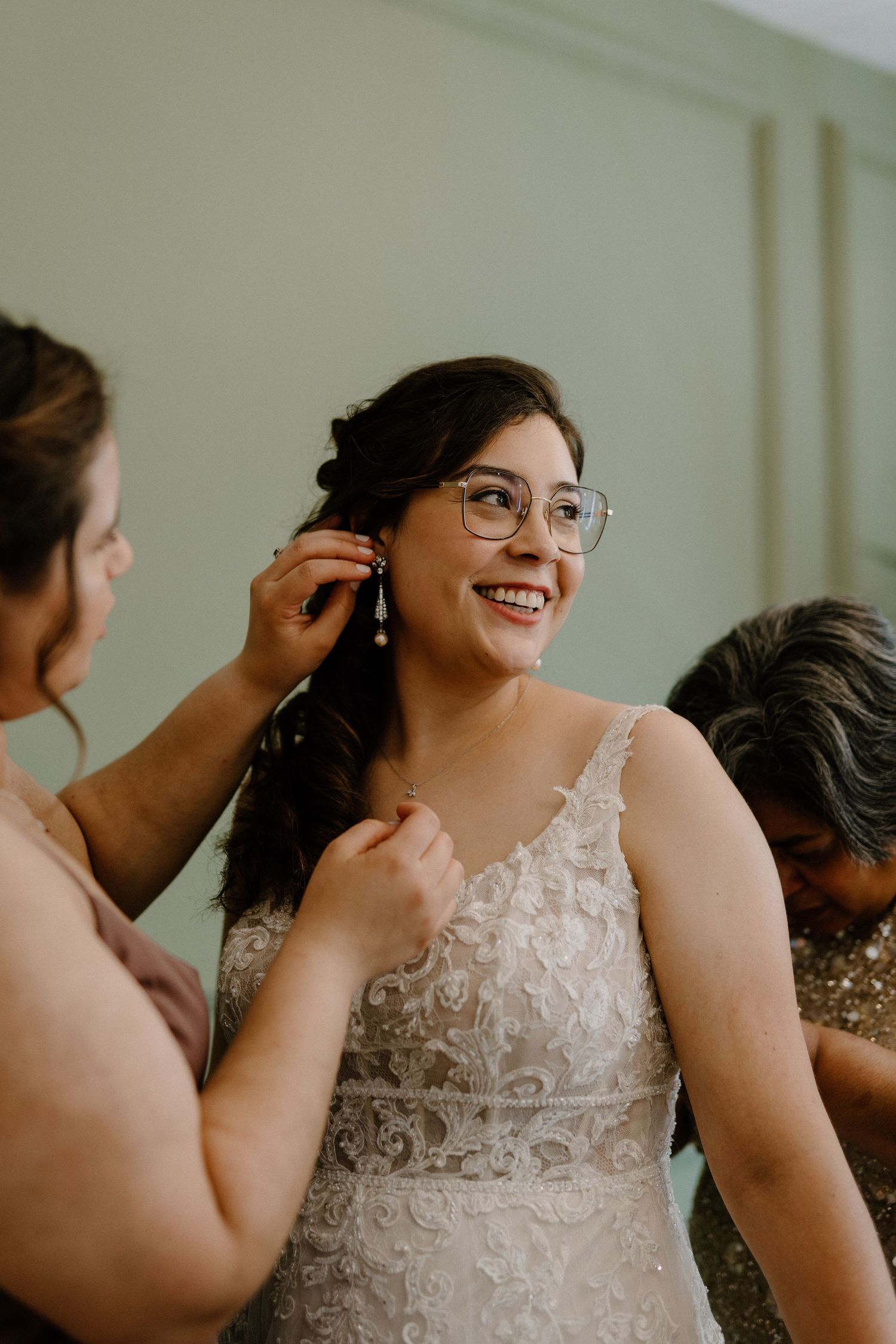 Smiling bride in lace wedding gown wearing glasses gets earrings put on by bridesmaid before ceremony.