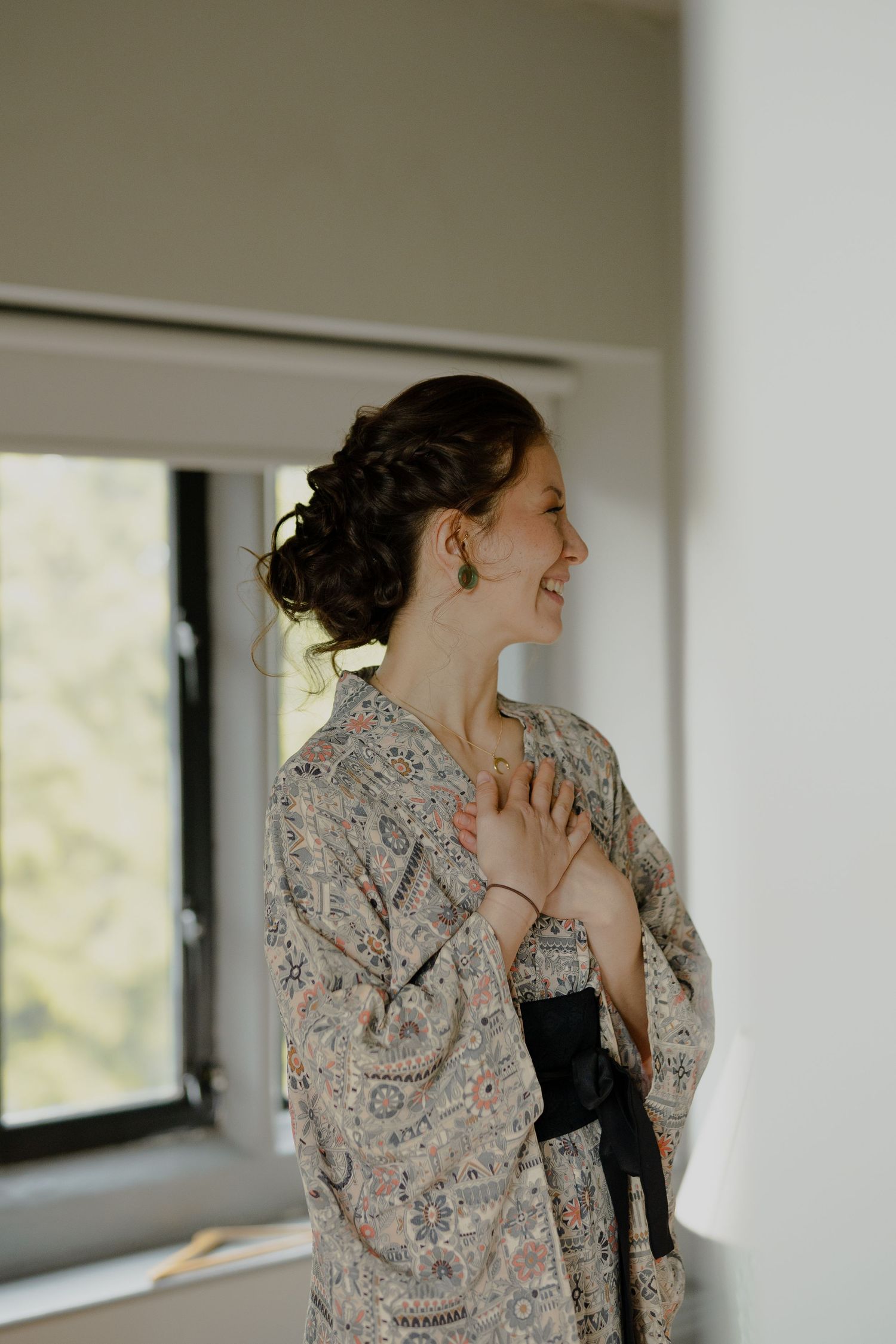 Happy woman in floral kimono robe laughing by a window during bridal wedding preparations.