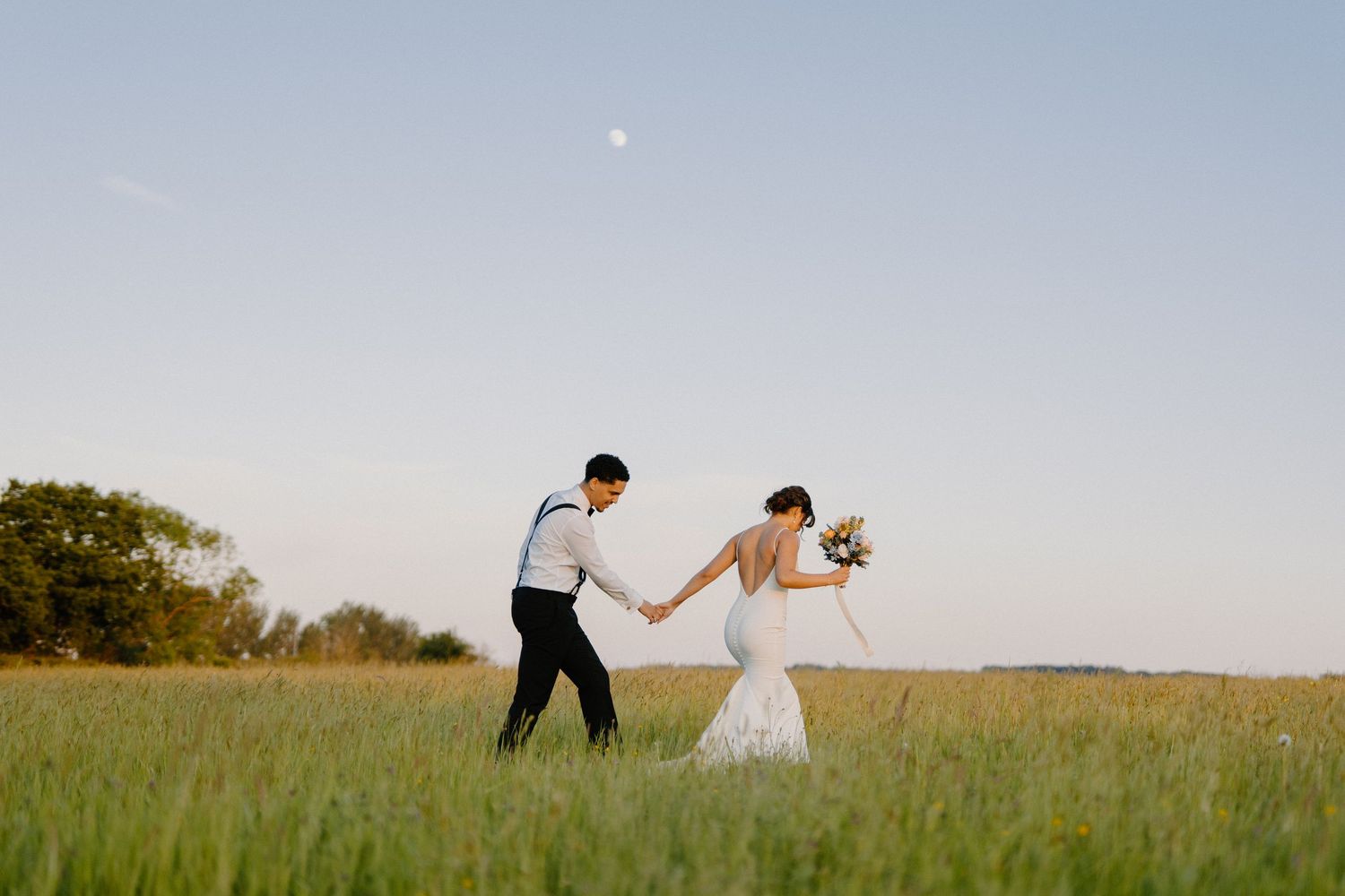 A bride and groom walk hand in hand through a lush green field under a clear blue sky with the moon visible.