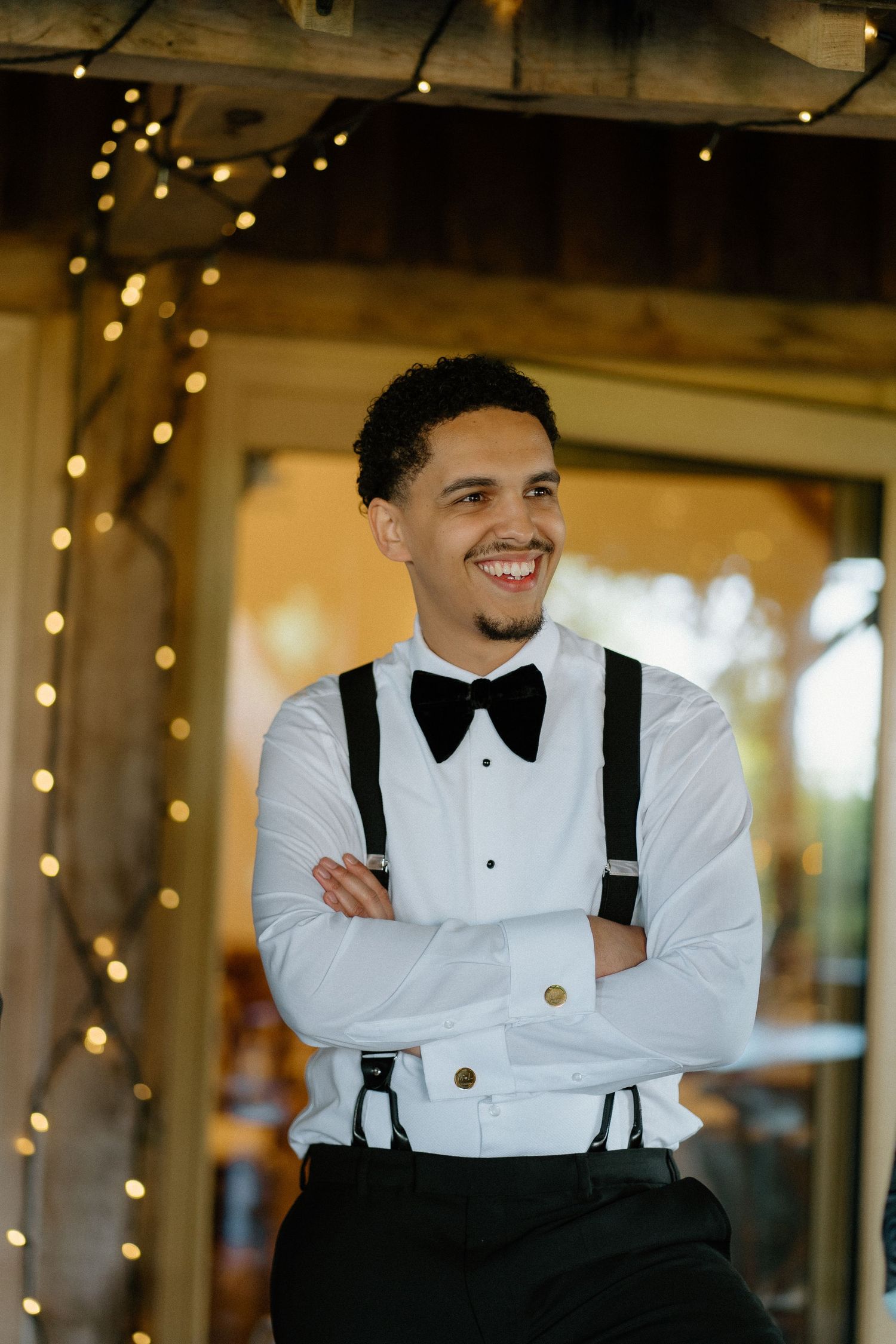 Smiling groom in white shirt, bow tie, and suspenders posing under warm string lights.