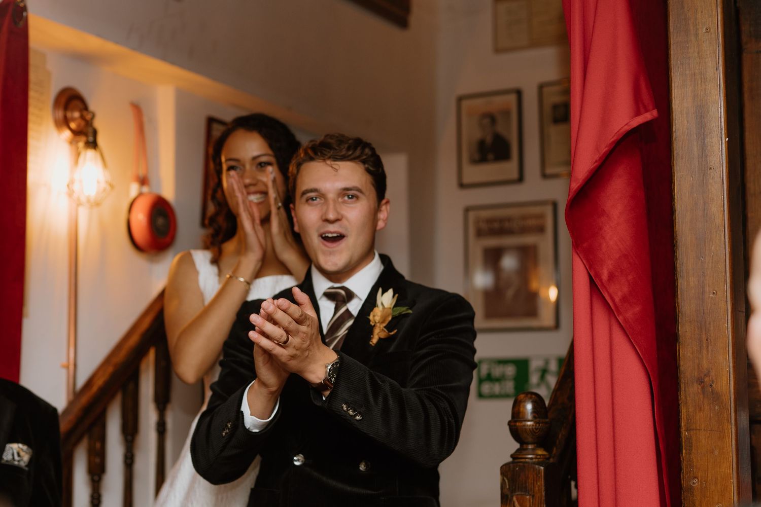 A groom in a dark suit claps and smiles with his bride on a staircase at their wedding reception.