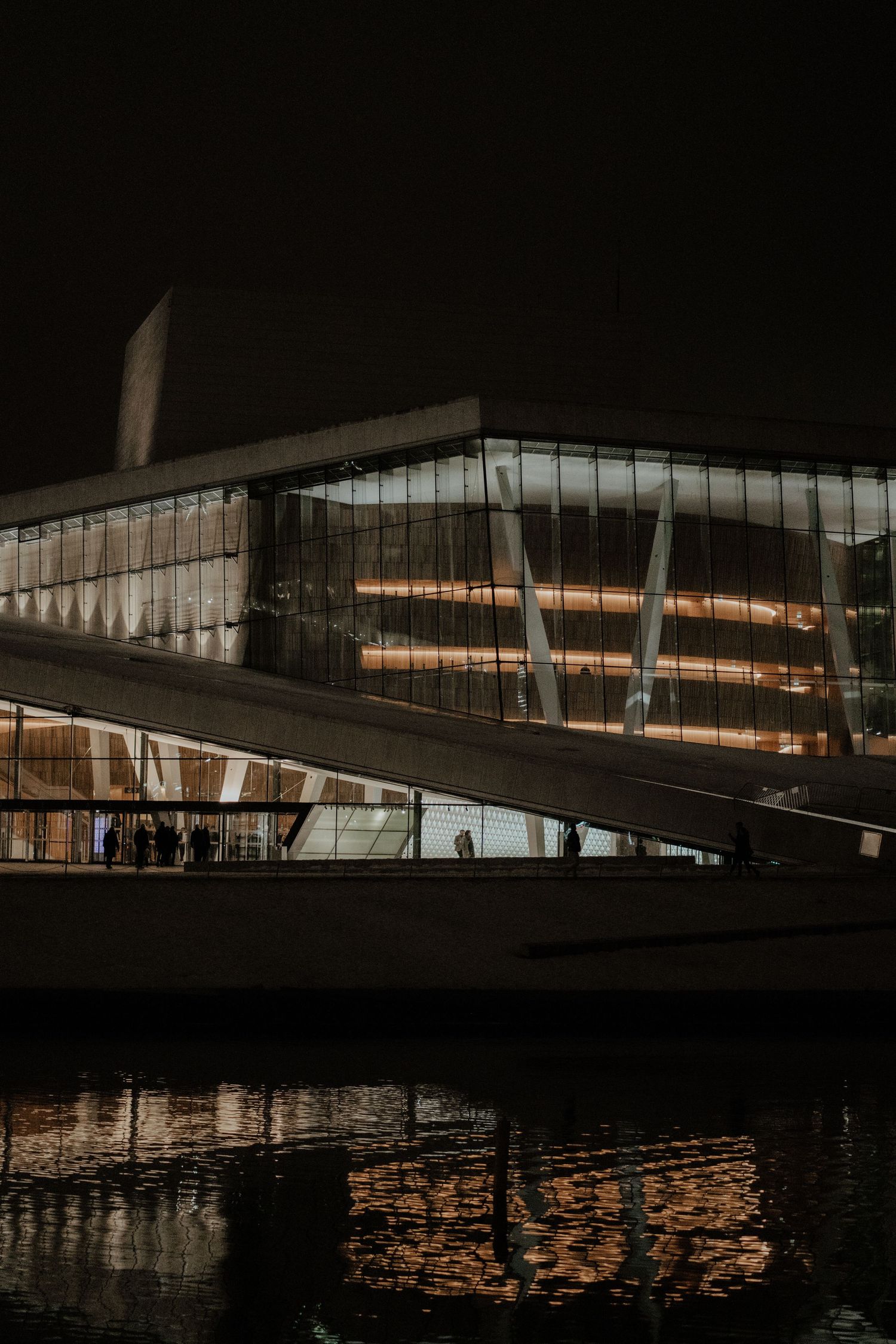 Modern glass building with illuminated levels reflected in dark water at night, featuring angular architectural design.