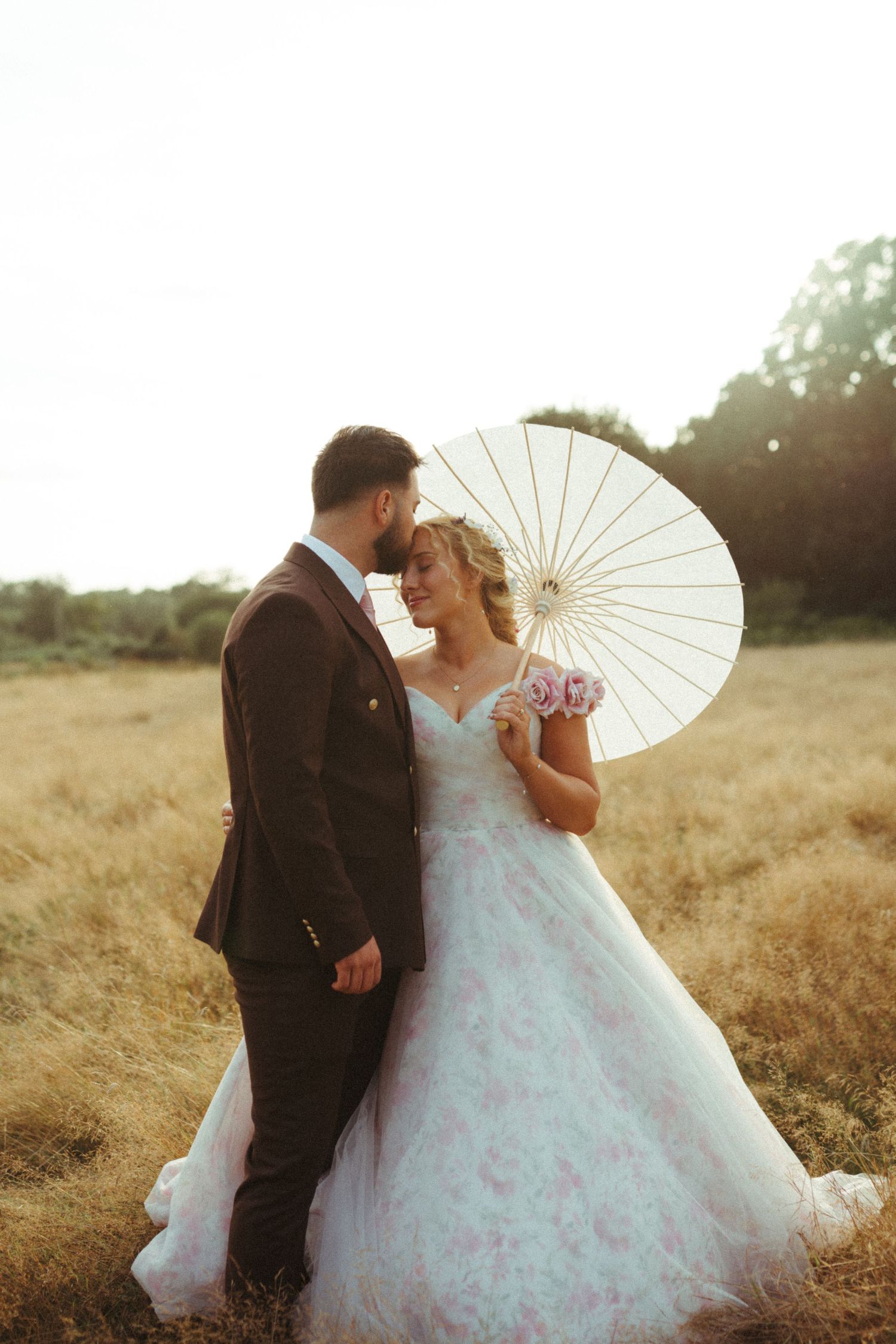 Newlyweds share an intimate moment with white parasol in golden field at sunset.