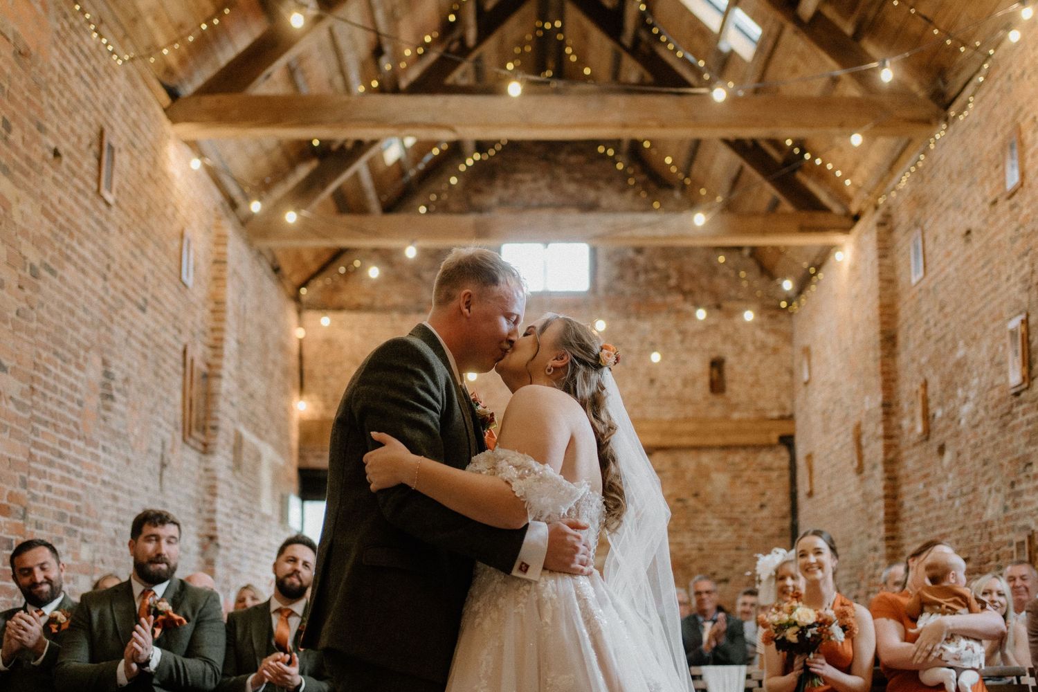 Bride and groom share first kiss at rustic barn wedding venue with fairy lights and exposed brick walls.