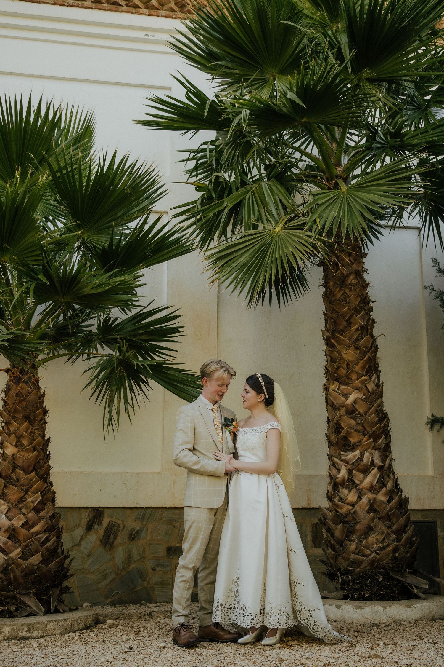 Bride and groom share a tender moment standing beneath tall palm trees at their wedding venue.