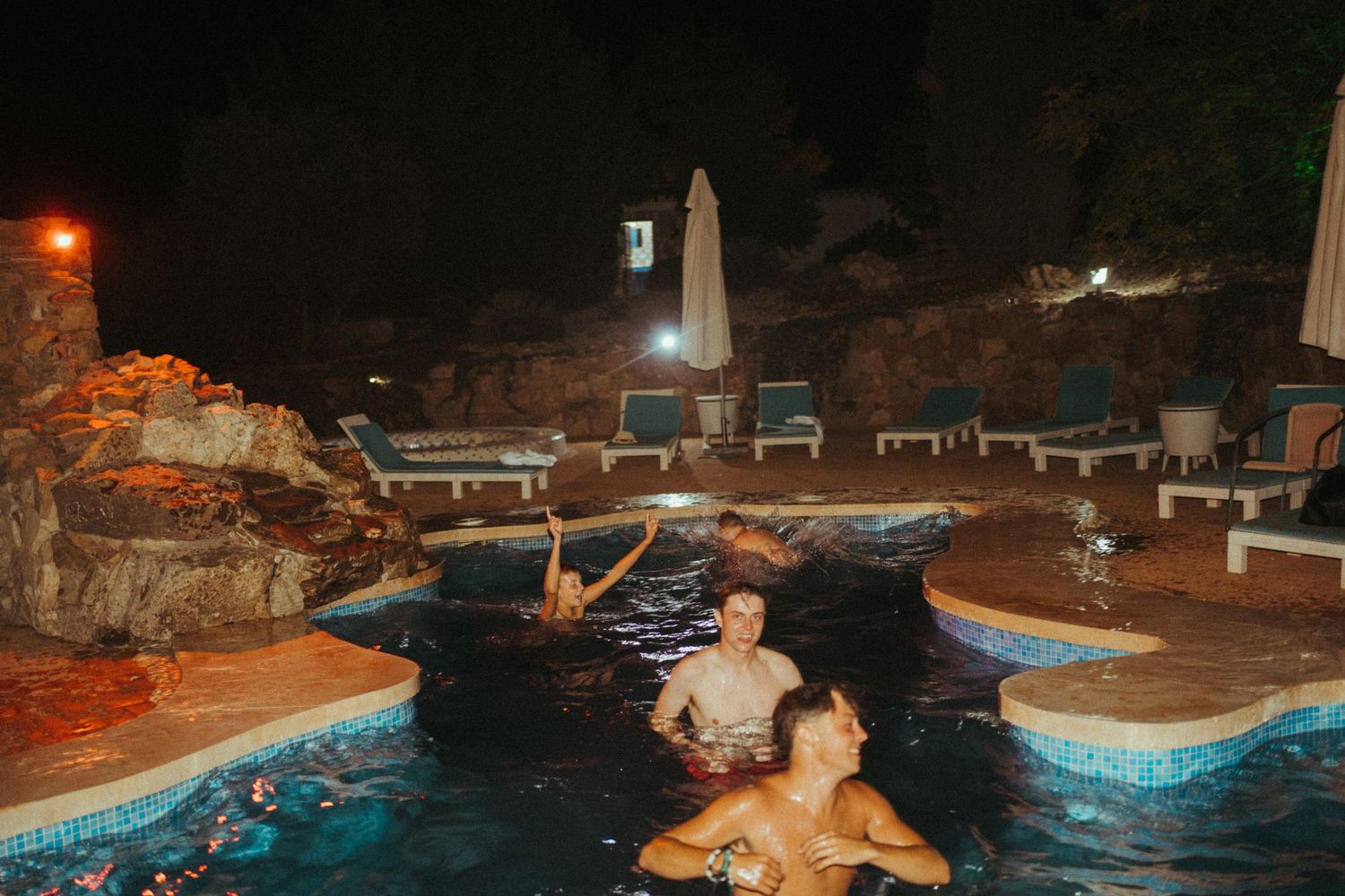 Guests laughing and swimming in an illuminated outdoor pool during a late-night wedding celebration.