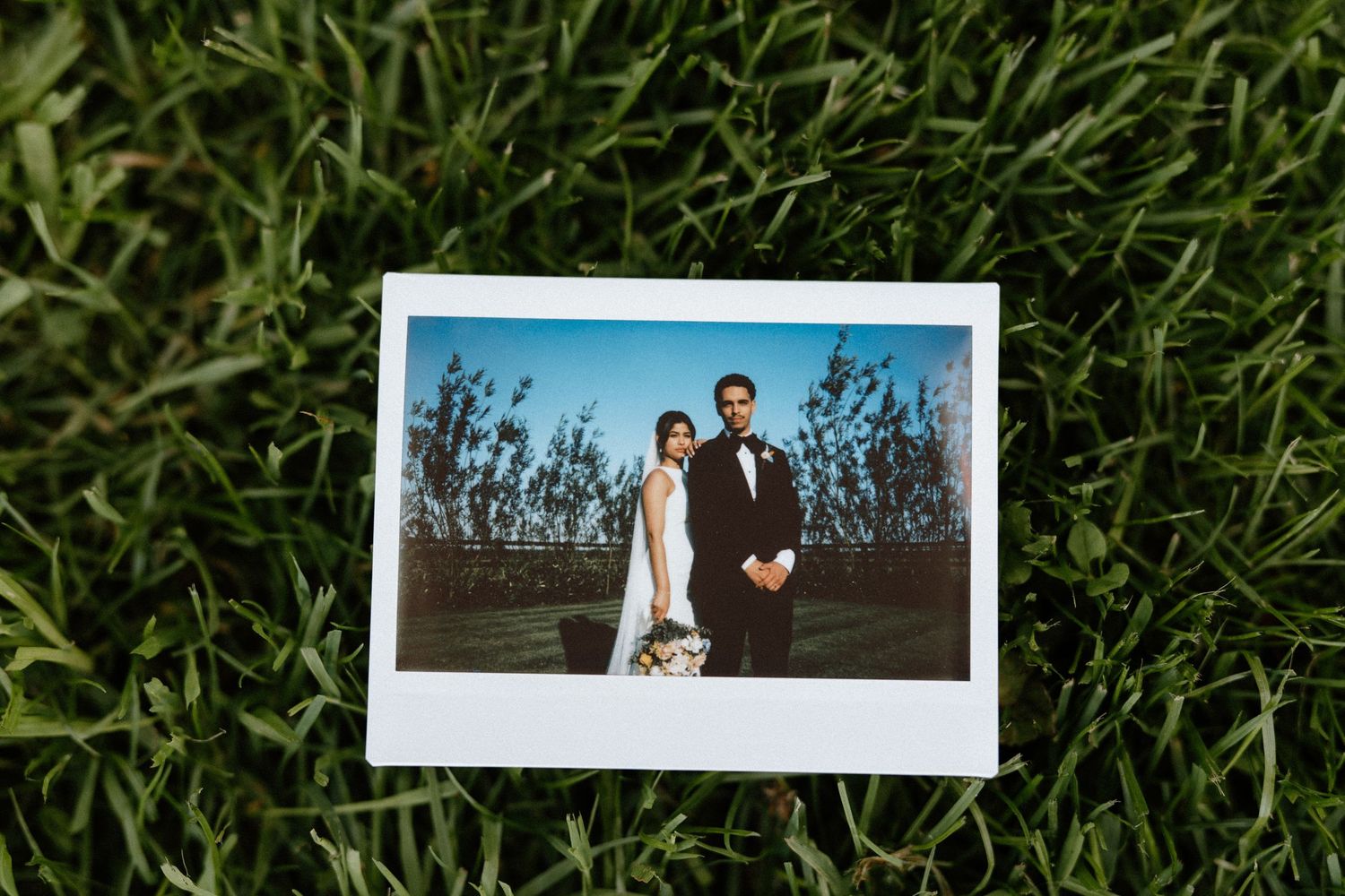 Polaroid photo of bride and groom standing together on green grass at outdoor wedding.