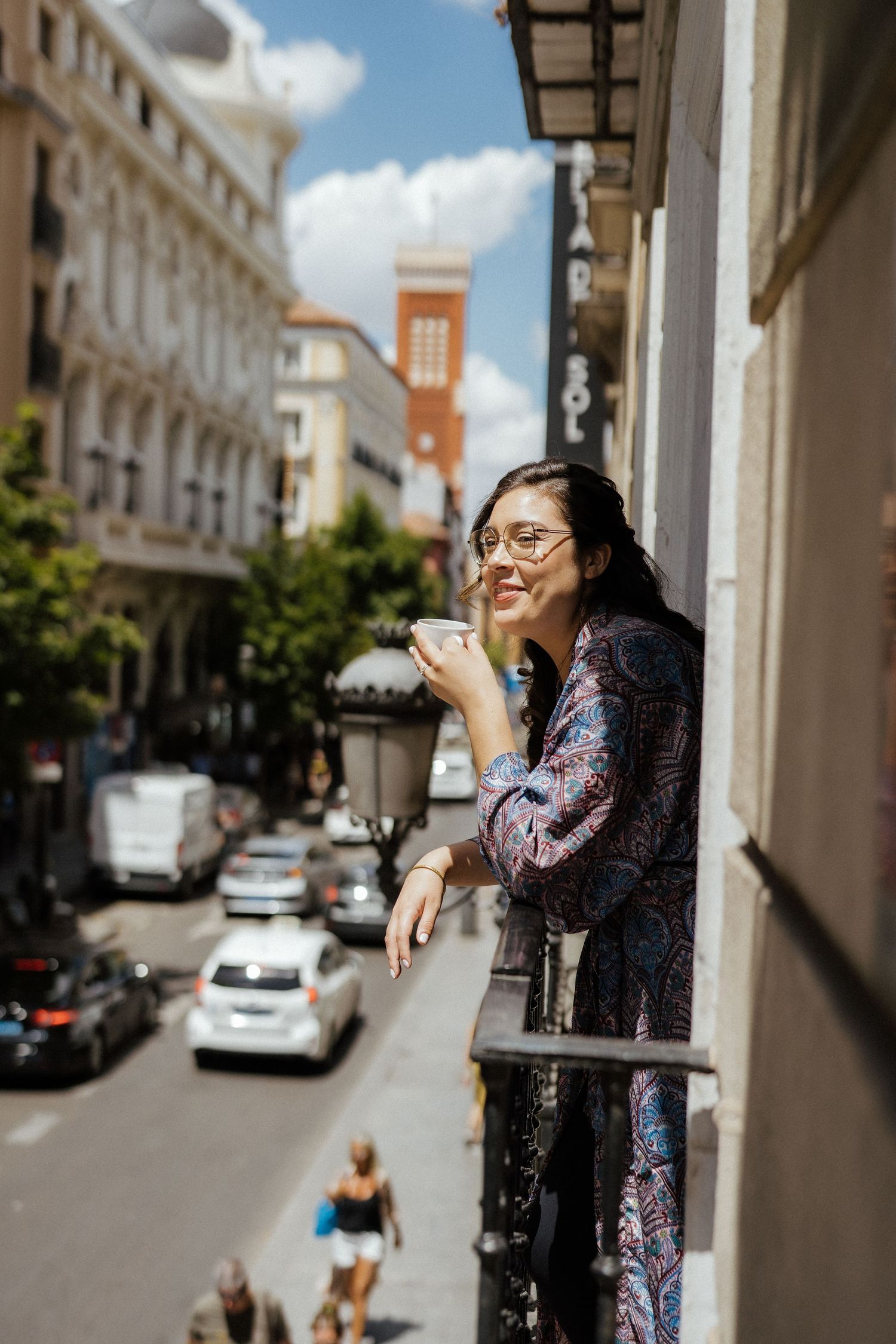 Smiling woman in floral dress enjoying coffee on a balcony with a vibrant city street view below.