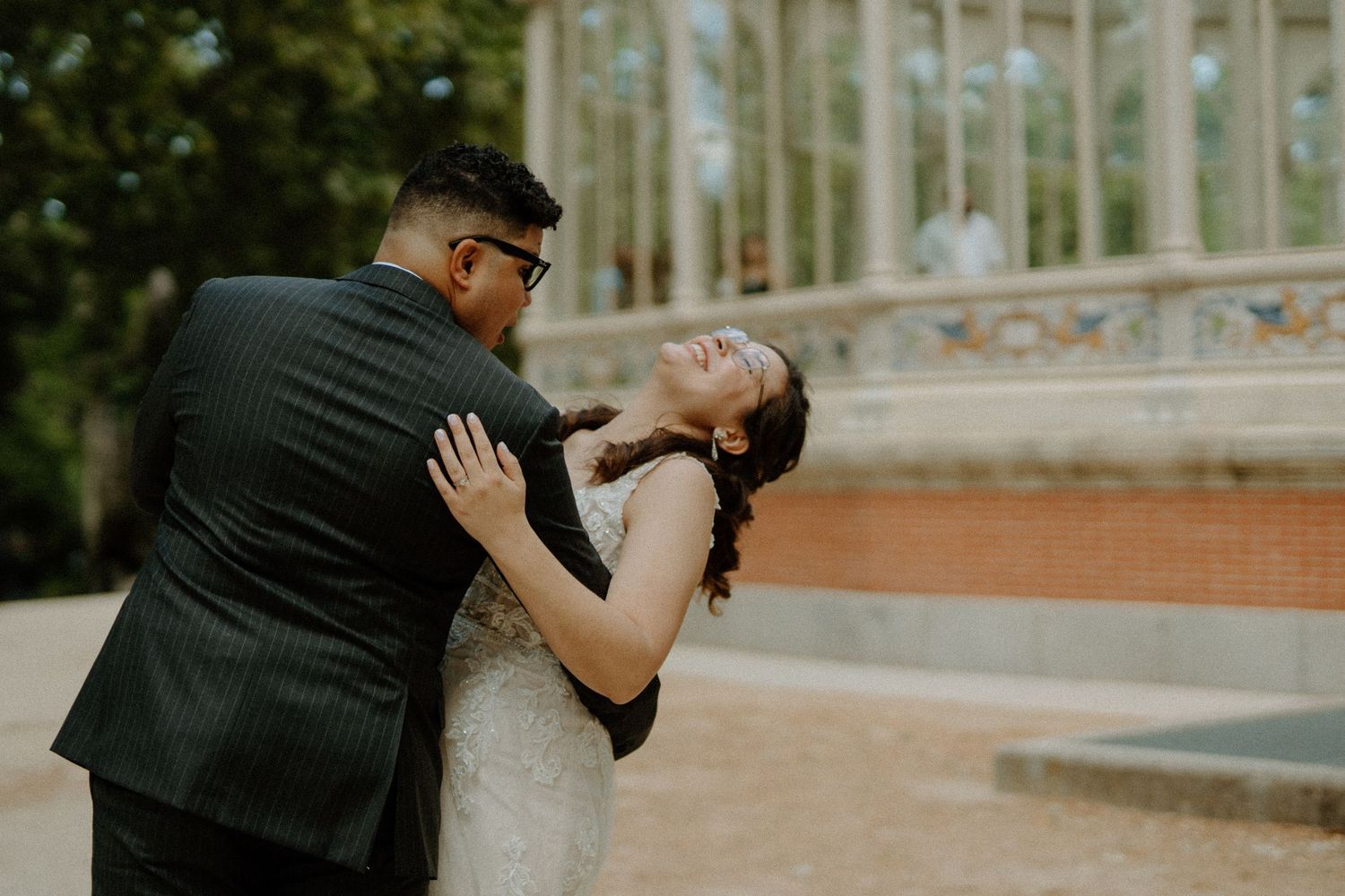 Groom tenderly holding bride outdoors near a decorative glass structure.