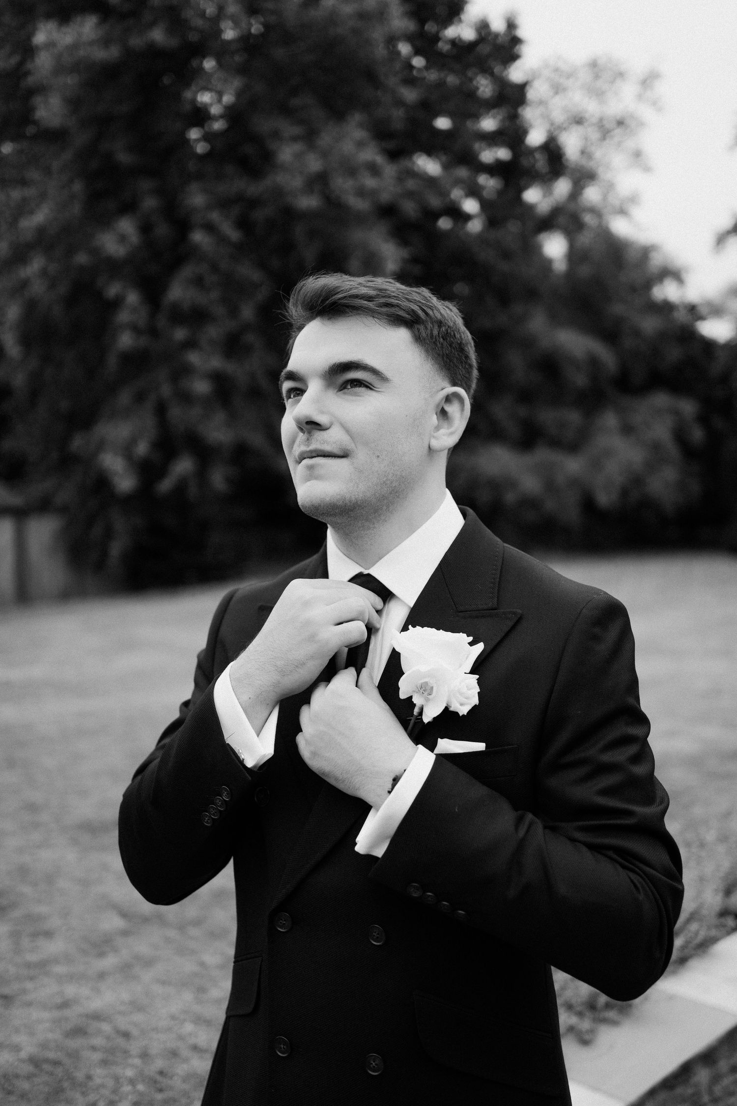 Groom in black suit adjusting tie with boutonniere, black and white outdoor portrait.