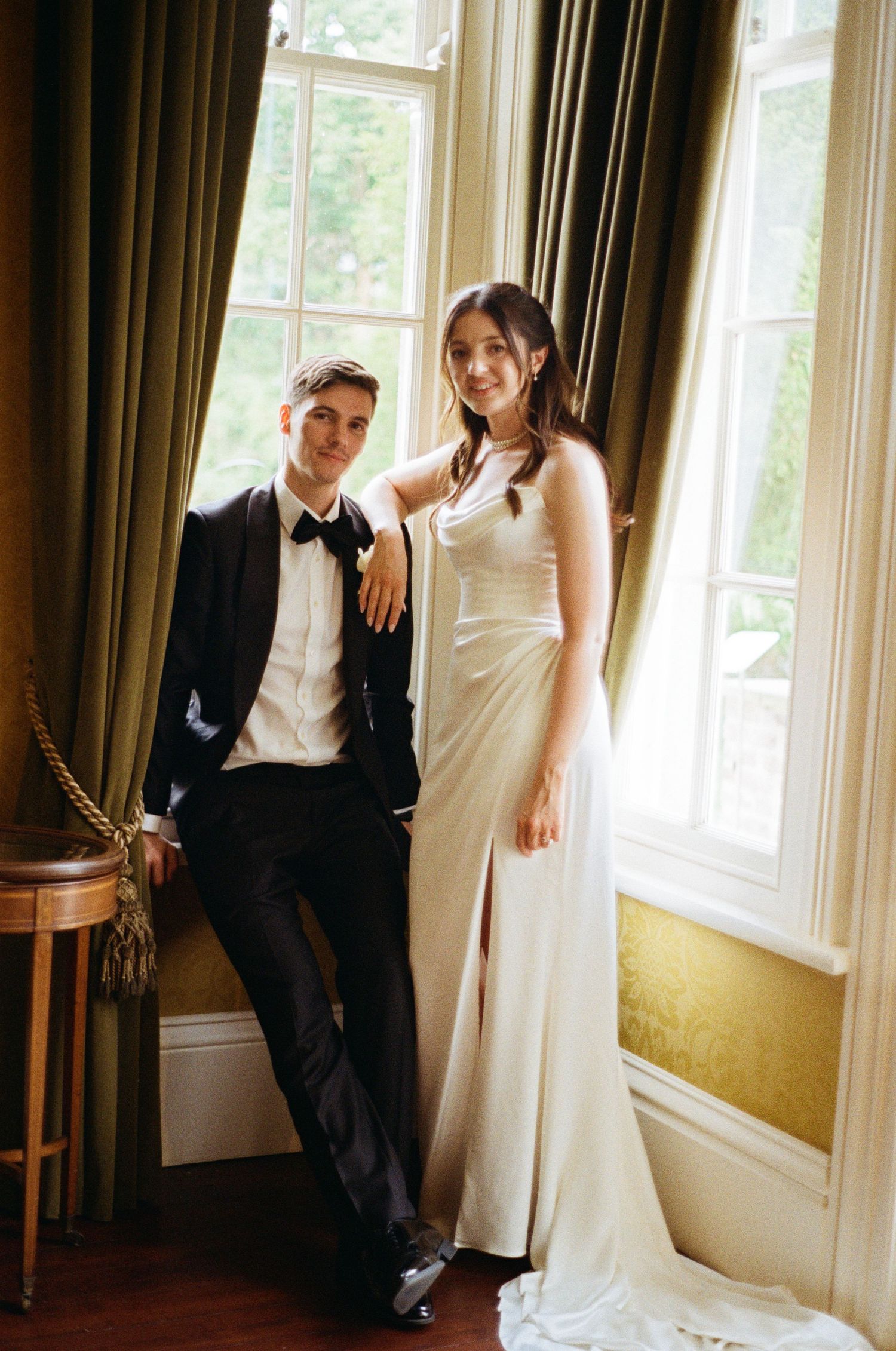 Bride and groom posing elegantly by tall green-draped windows inside a classic country house venue.